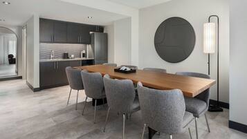 Een moderne eetruimte met een houten tafel en zes grijs beklede stoelen. Op de muur hangt een groot zwart rond kunstwerk, en rechts is een vloerlamp zichtbaar.