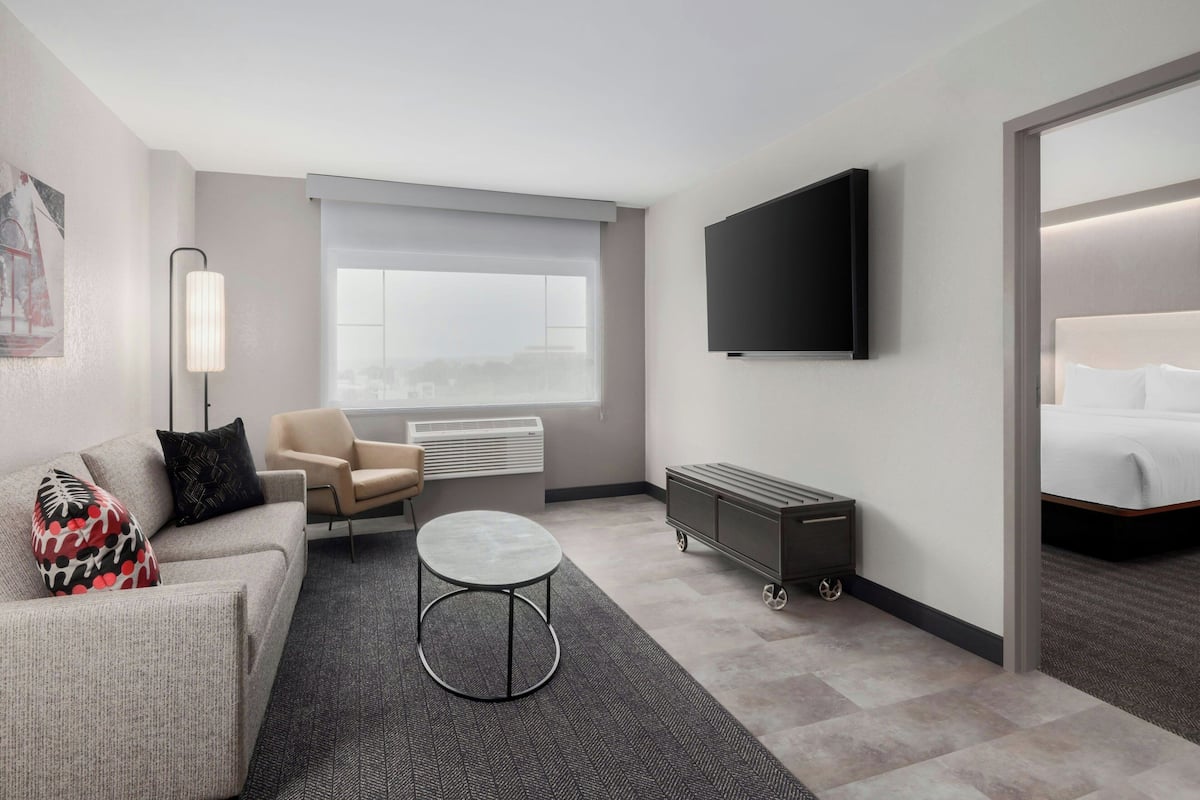 A modern hotel room with a sofa, armchair, round coffee table, TV, and a bed.