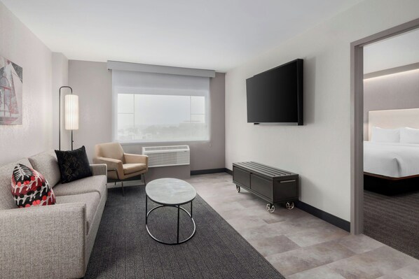 A modern hotel room with a sofa, armchair, round coffee table, TV, and a bed.