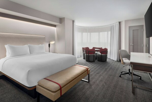 A hotel room with a large bed, a desk, a chair, a bench, and a dining area with chairs and a table. - Courtyard by Marriott Athens Downtown (Athens)