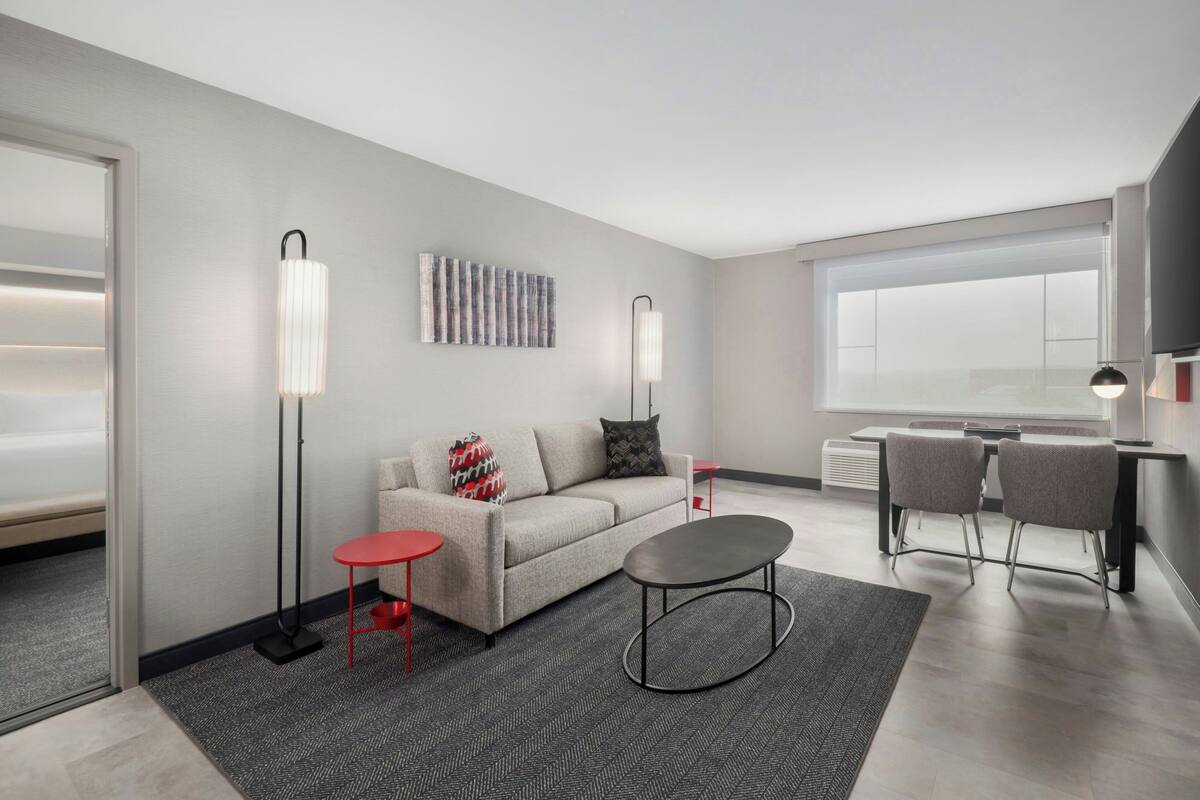 A modern living room with a sofa, a coffee table, a red side table, and a floor lamp. There is a large window with blinds, a flat-screen TV mounted on the wall, and a decorative wall piece above the sofa.