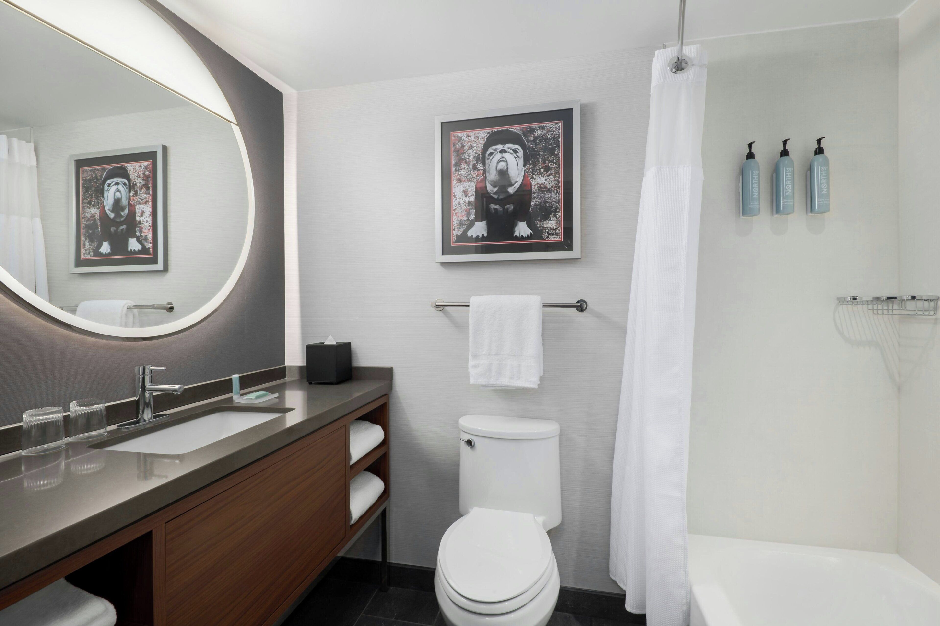 A modern bathroom with a sink, toilet, and shower. There are framed pictures on the wall, a towel rack, and toiletries on the wall.