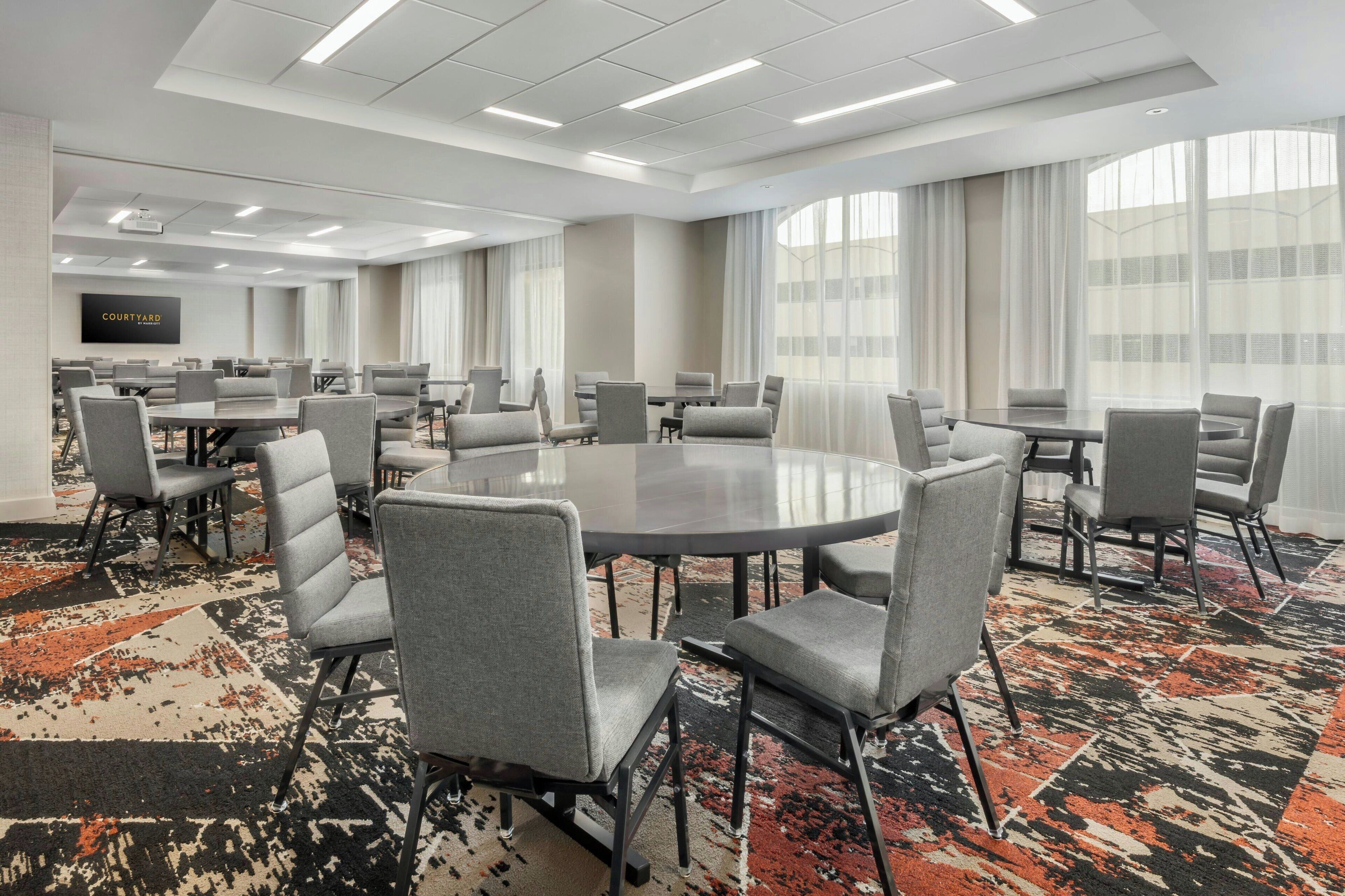 A conference room with round tables and grey chairs, large windows with curtains, and a carpeted floor.