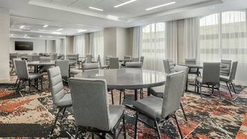 A conference room with round tables and grey chairs, large windows with curtains, and a carpeted floor.