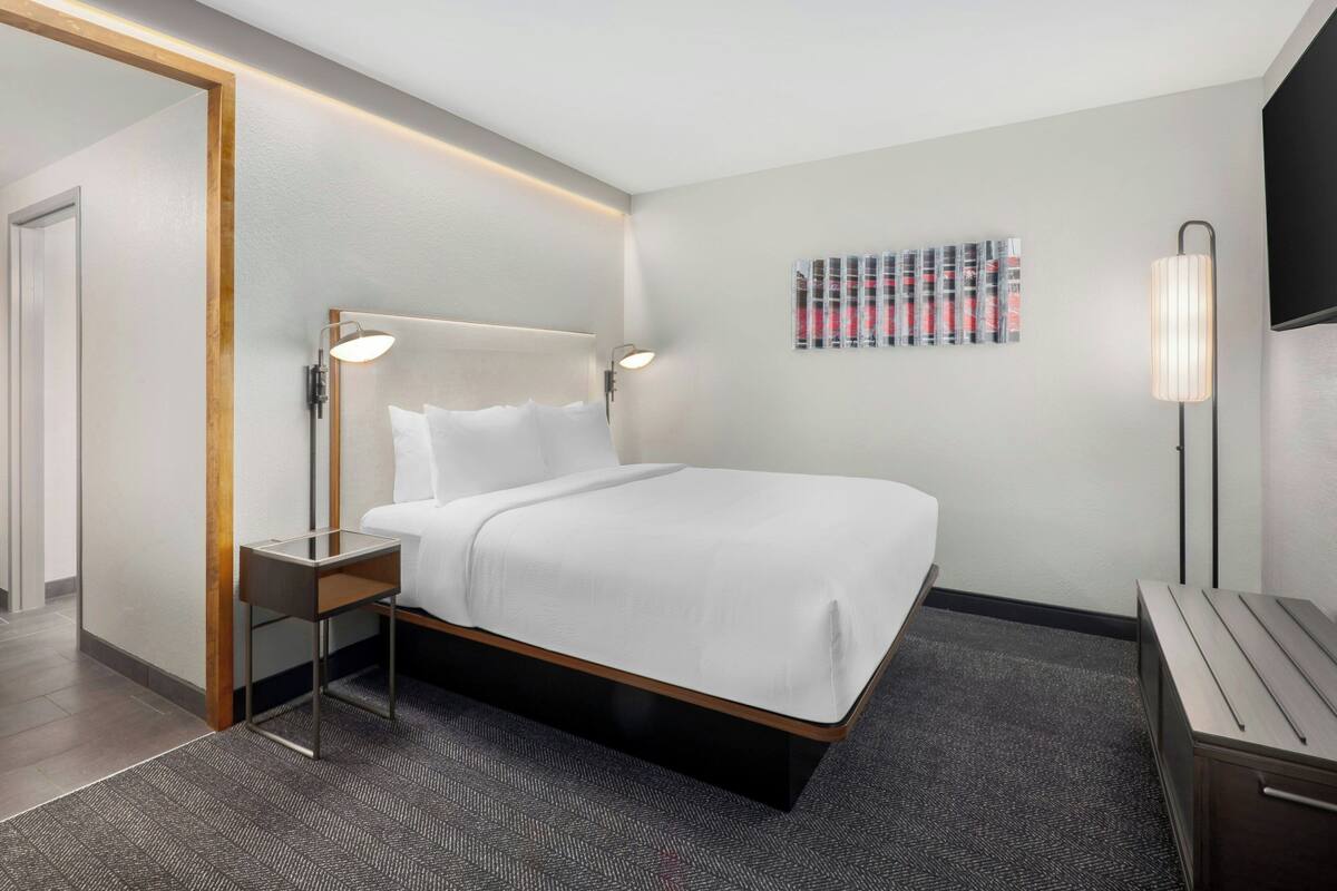 A modern hotel room with a neatly made bed, a bedside table with a lamp, a wall-mounted TV, and a carpeted floor.