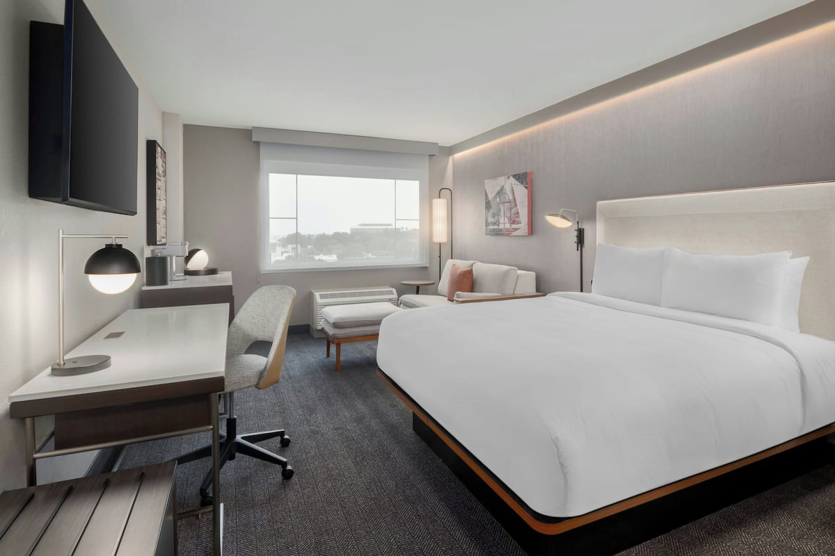 A modern hotel room with a large bed, a desk with a chair, a flat-screen TV, a window, and a wall-mounted lamp.