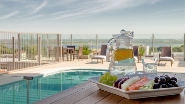 Una mesa con una jarrón de agua, un vaso de agua y un plato de fruta junto a una piscina con vistas al océano.