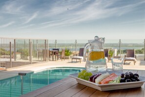 Una mesa con una jarrón de agua, un vaso de agua y un plato de fruta junto a una piscina con vistas al océano.