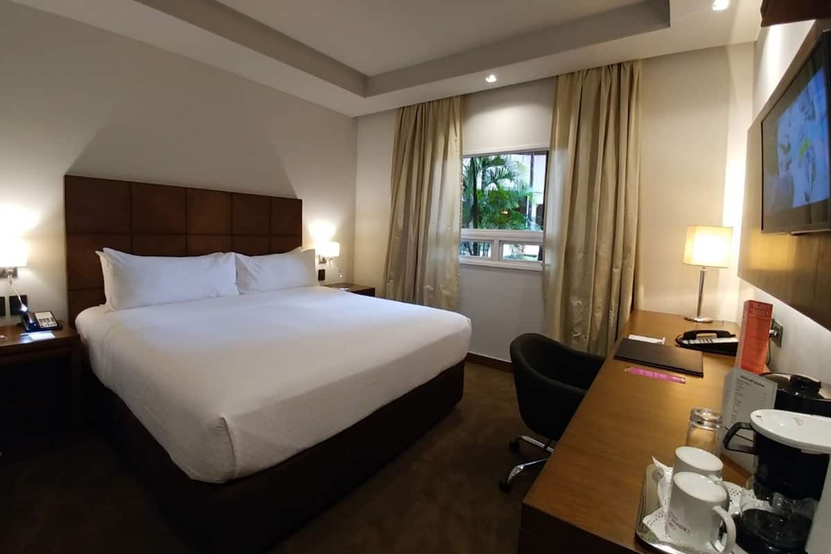 A hotel room with a large bed, a desk with a coffee maker, a chair, a TV, and a window with curtains.