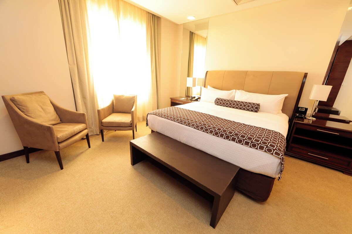A hotel room with a bed, a bench, two armchairs, a nightstand, and a mirror.