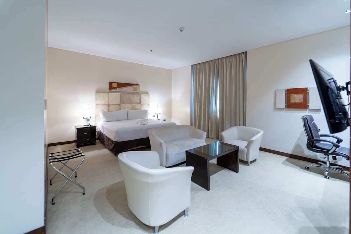 A hotel room with a bed, two white armchairs, a small table, a desk, a chair, and a TV.