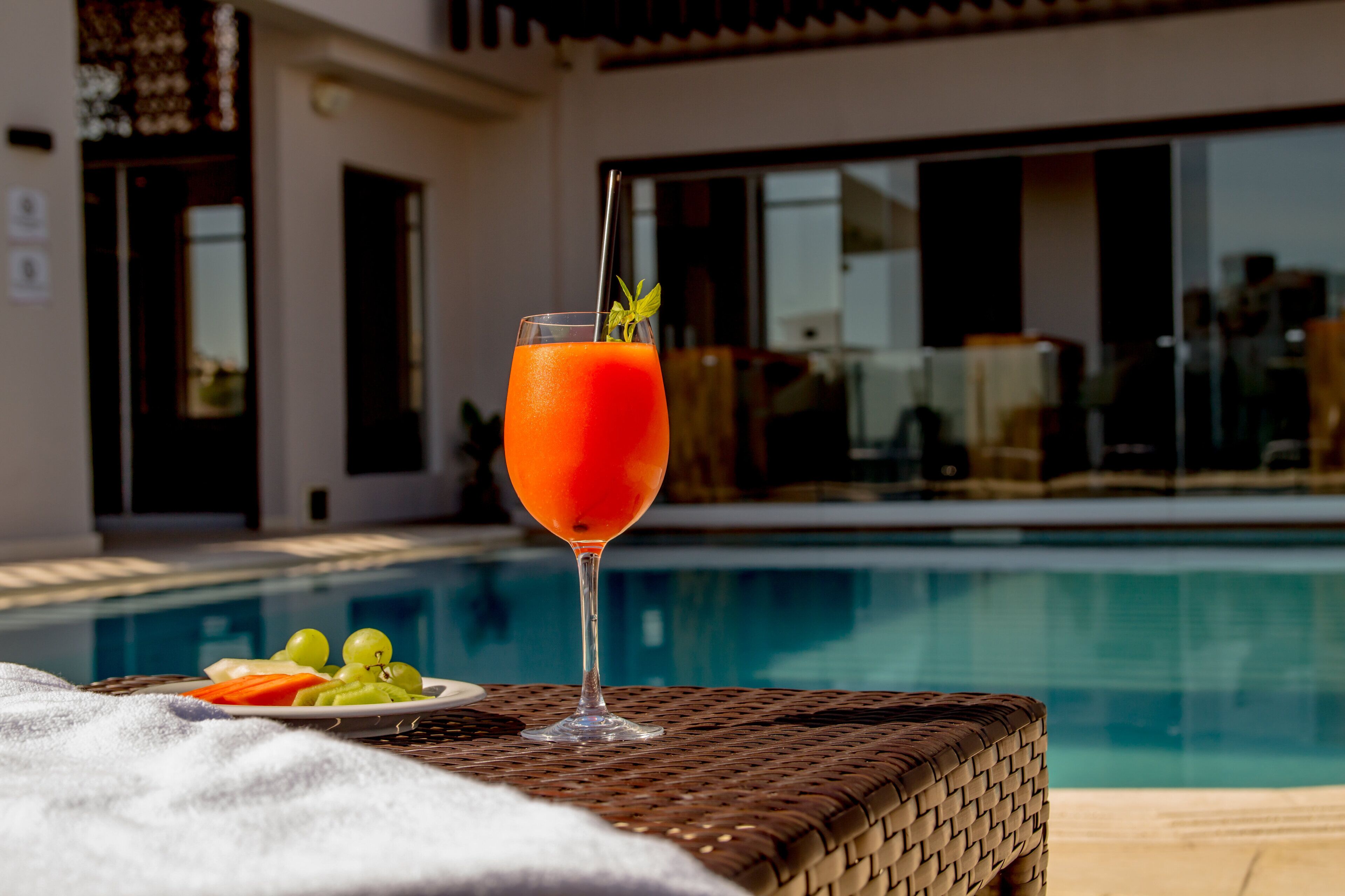 A glass of orange drink with a straw and mint on a wicker table by a pool.