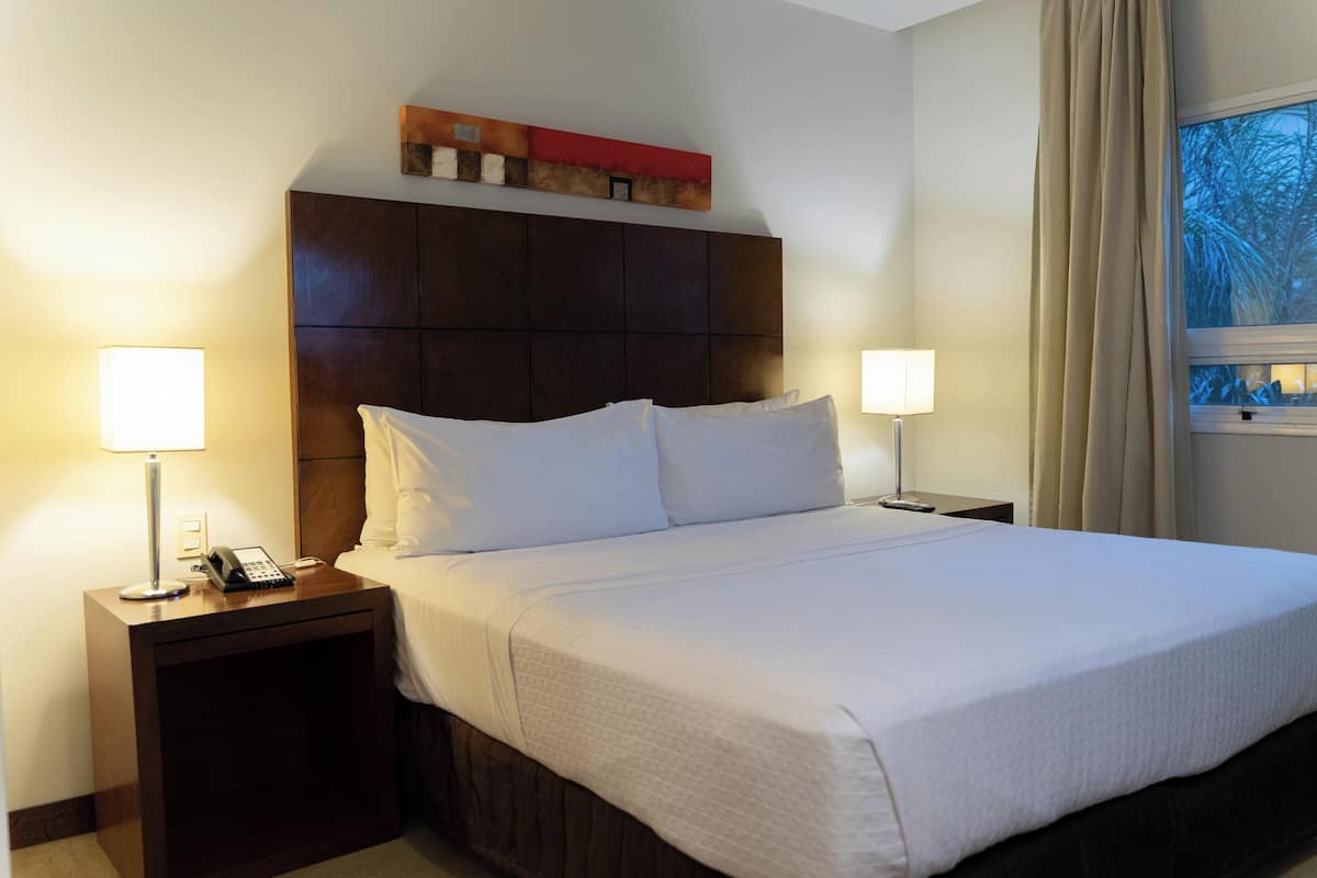 A hotel room with a large bed, two bedside tables with lamps, a wooden headboard, and a window with curtains.