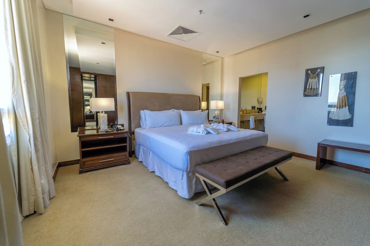 A hotel room with a large bed, bedside tables, a bench, a mirror, and artwork on the walls.