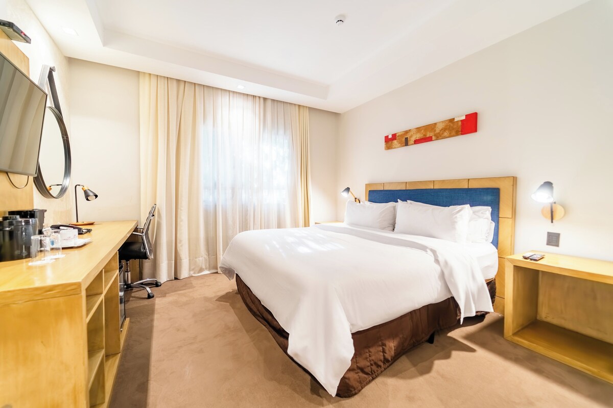 Habitación de hotel con cama, escritorio, silla y ventana con cortinas.