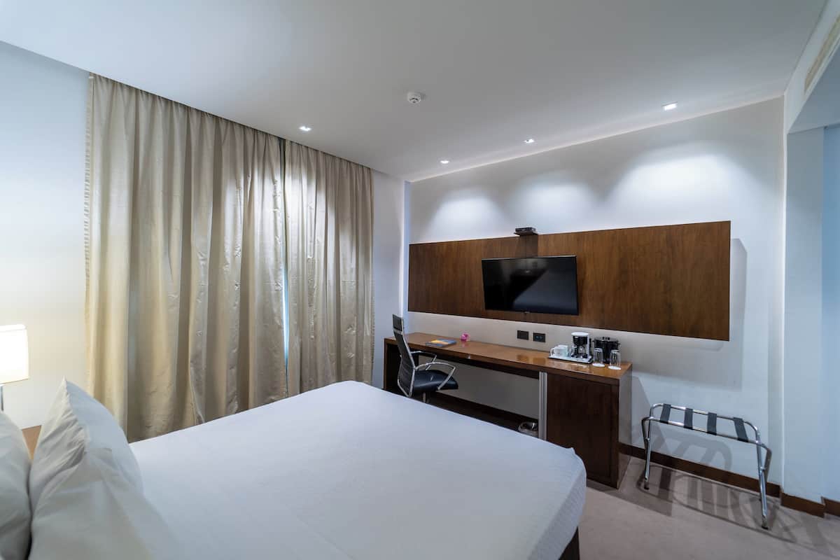 A hotel room with a bed, a desk with a computer, a TV mounted on the wall, and a window with curtains.