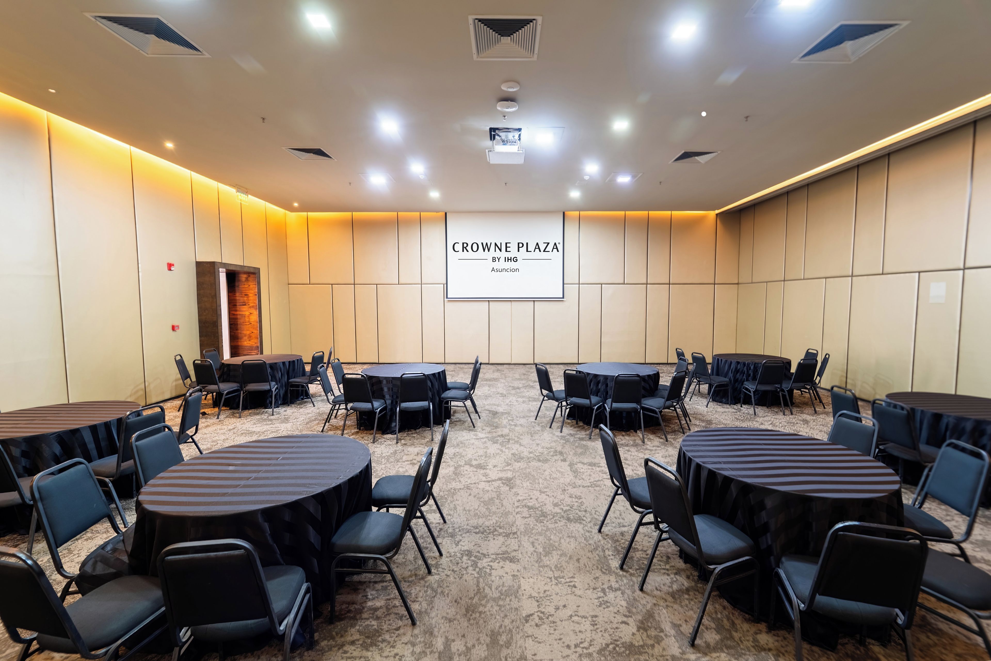 A conference room with multiple round tables and chairs, a projector screen displaying 'CROWNE PLAZA BY IHG', and a carpeted floor.