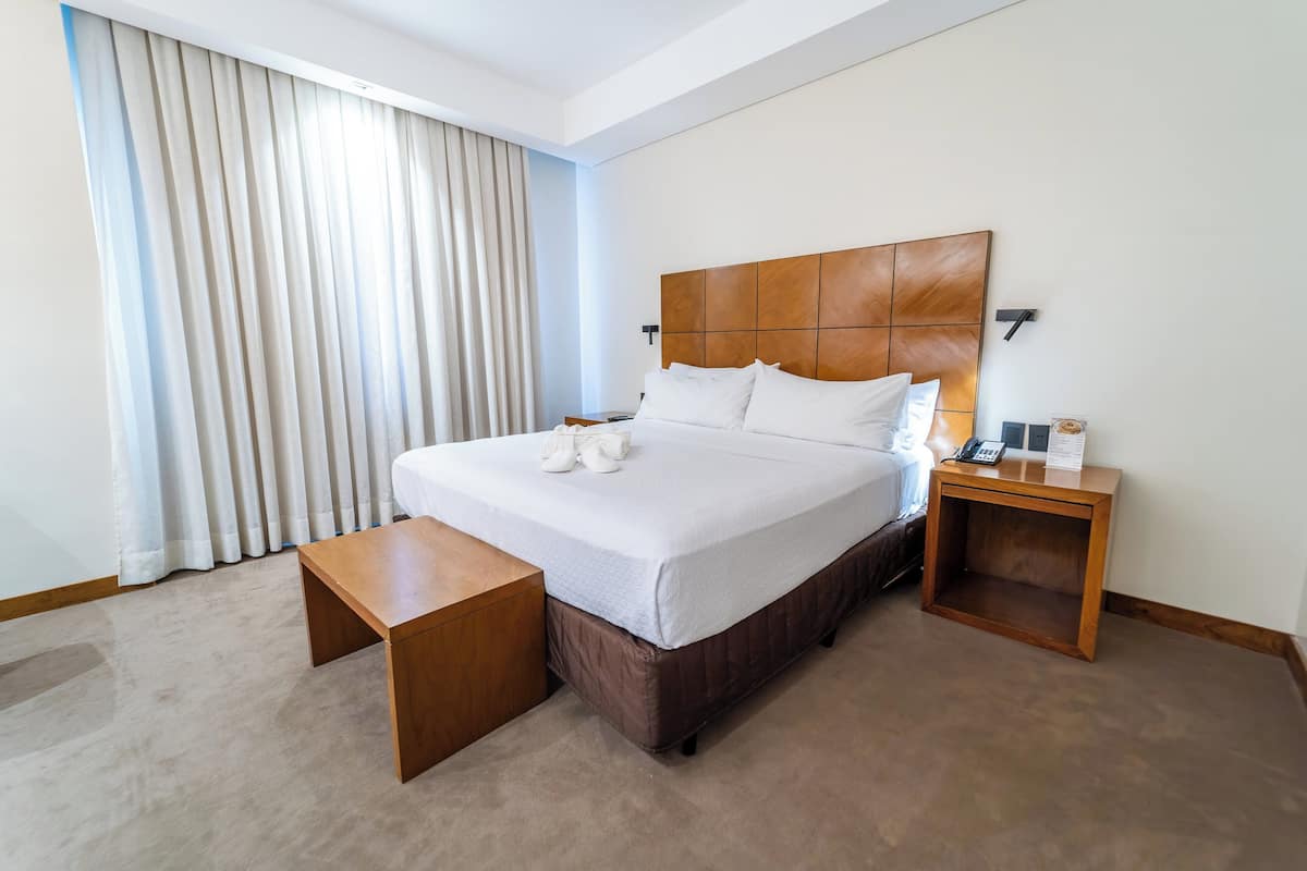 A hotel room with a large bed, a wooden nightstand, a bedside table, and a window with curtains.
