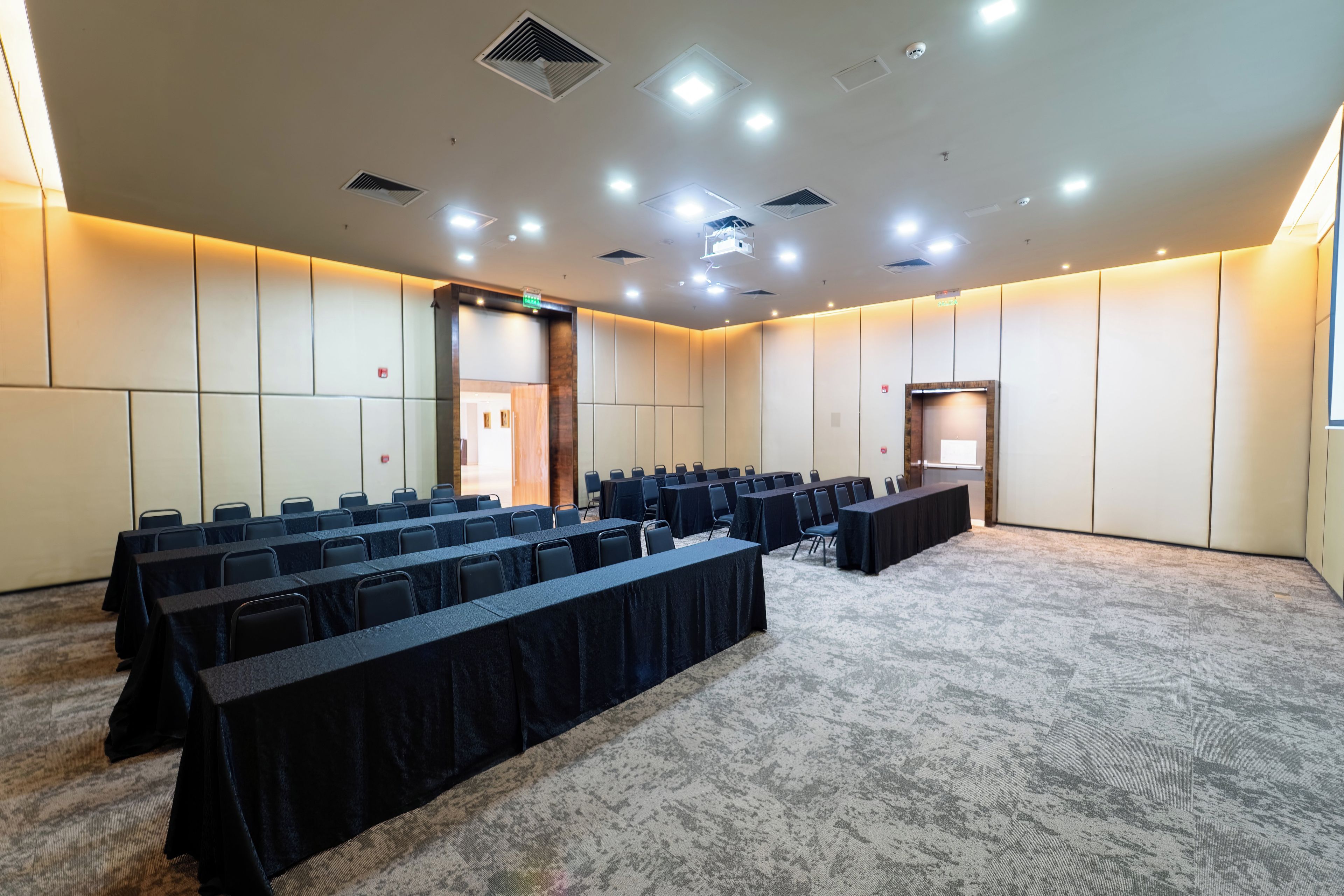 A conference room with rows of tables and chairs, a carpeted floor, and a high ceiling with recessed lighting.