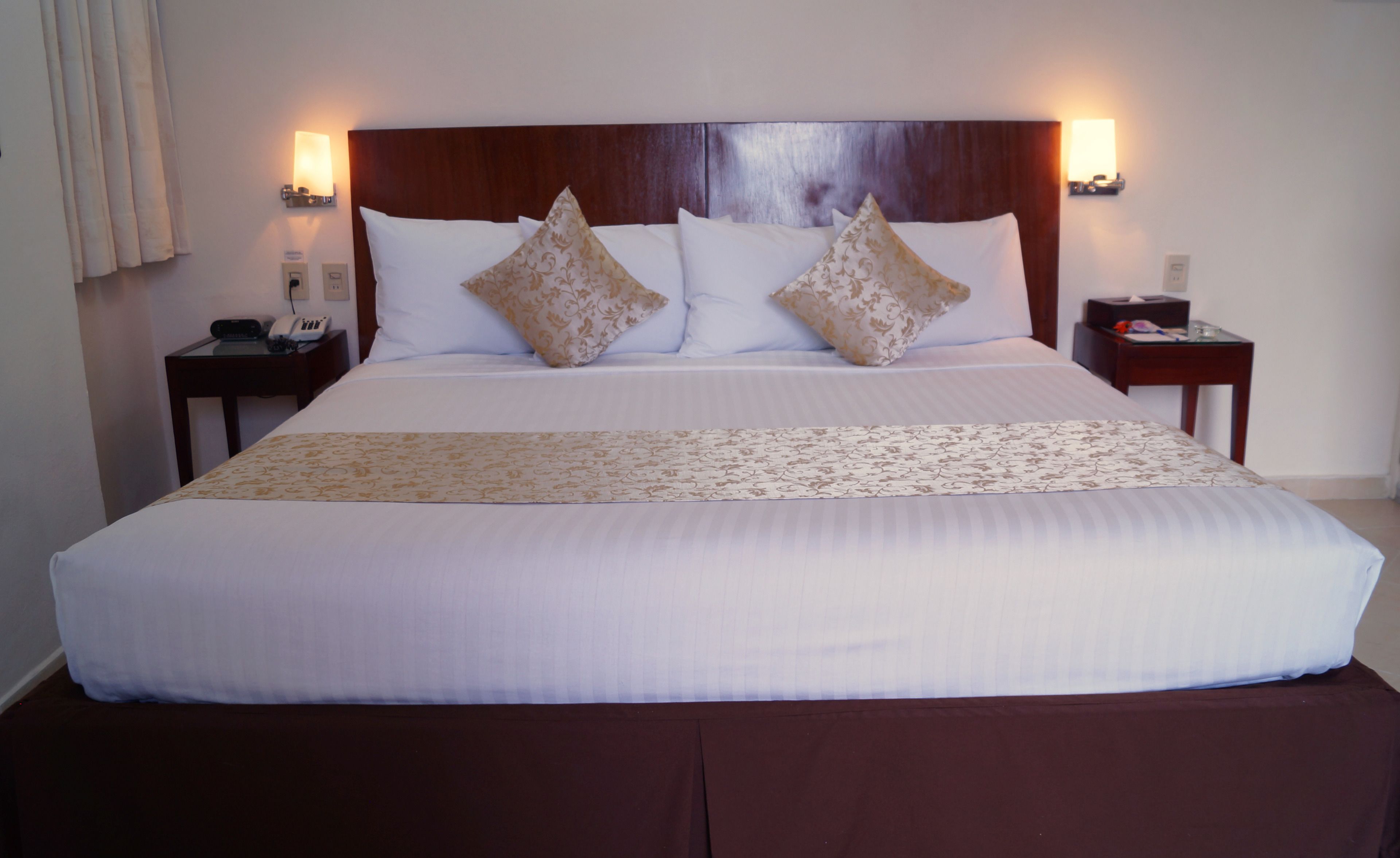 Una cama bien hecha con cabecera de madera, dos mesitas de noche con lámparas y una colcha estampada.