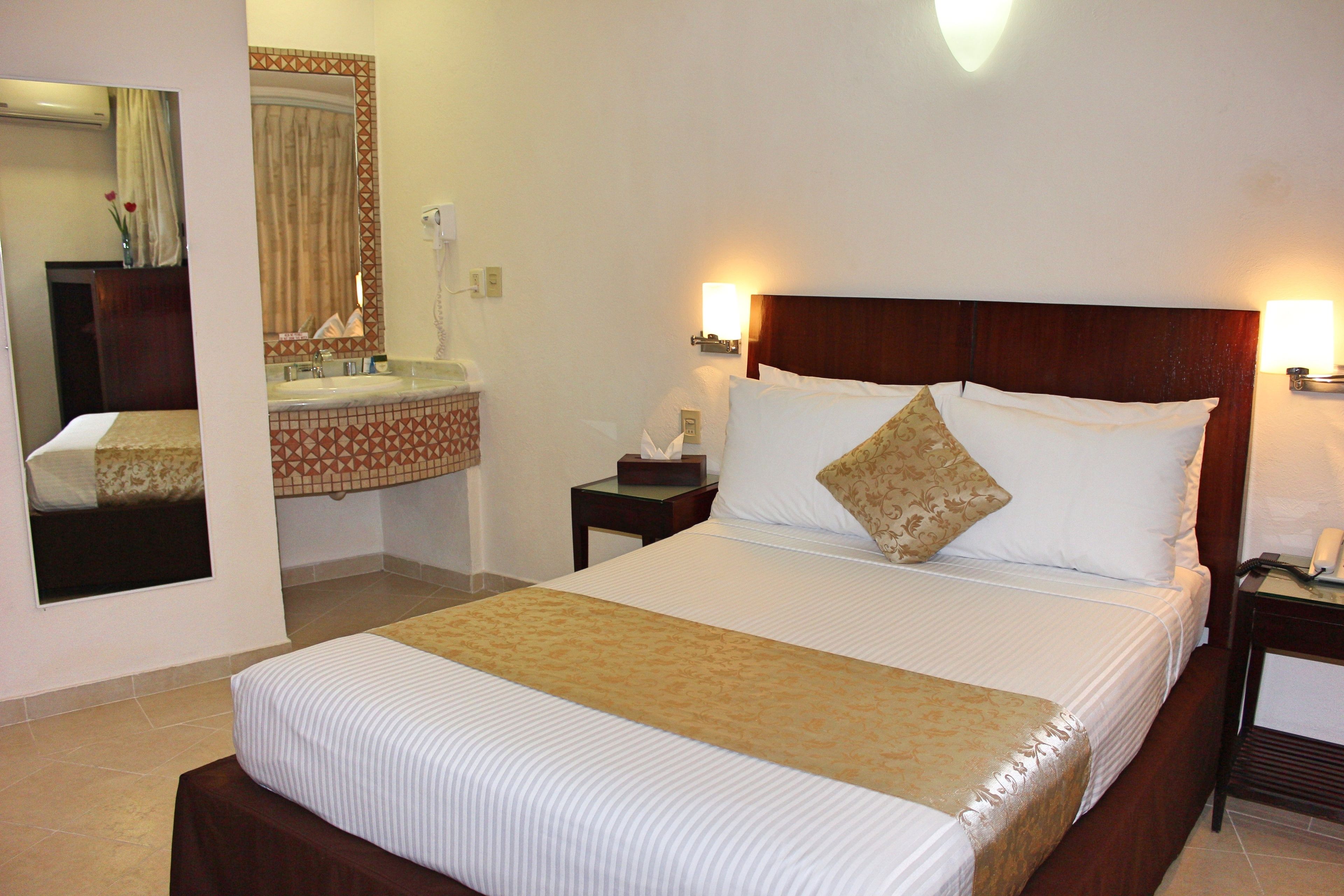 A hotel room with a large bed, a bedside table with a lamp, a nightstand, a mirror, and a bathroom with a sink and toilet.