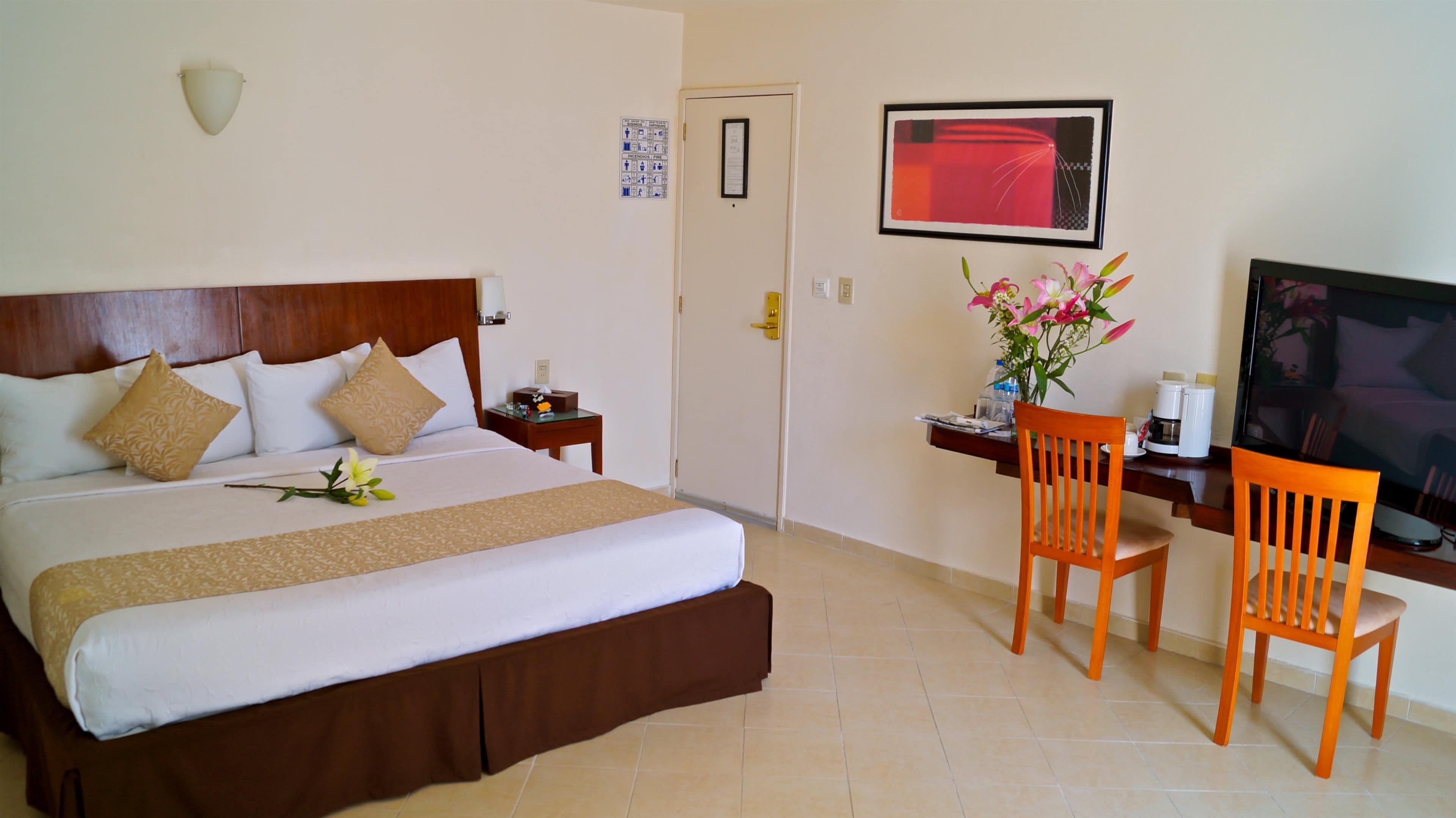 A hotel room with a bed, a desk with two chairs, a television, and a vase with flowers.