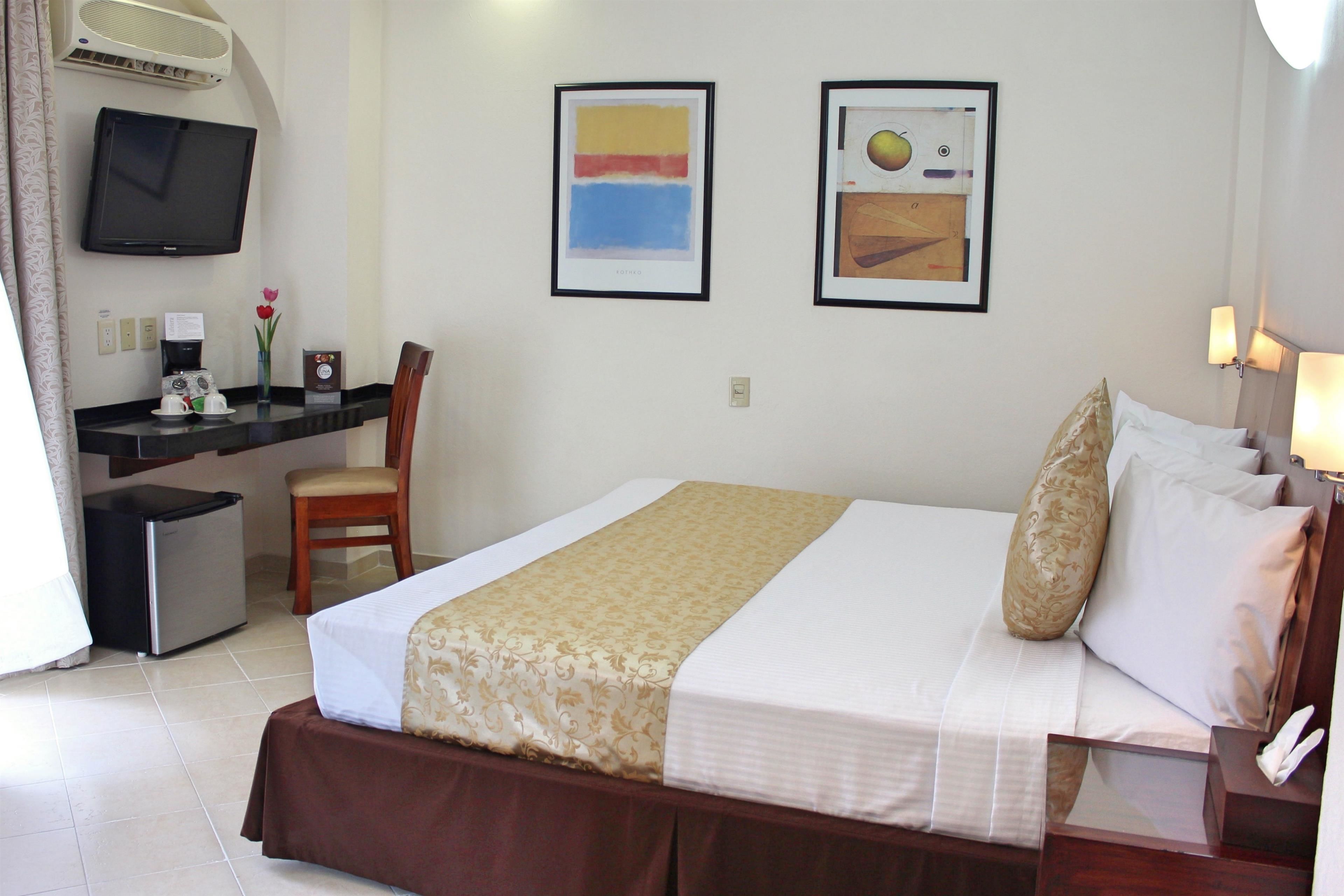 Habitación de hotel con cama, escritorio, silla, televisor y una obra de arte en la pared.