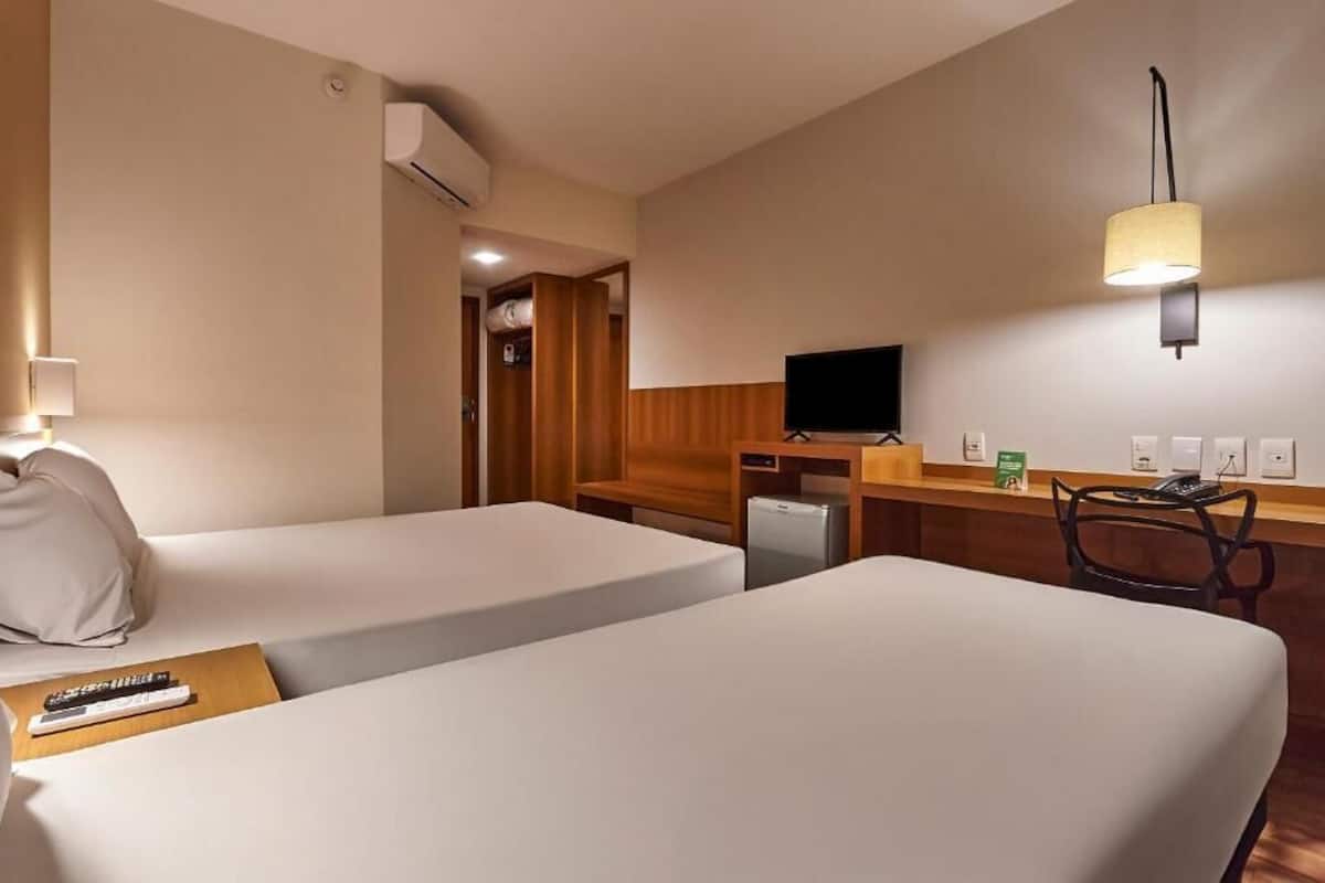 A hotel room with two beds, a desk, a television, a lamp, and a small refrigerator.