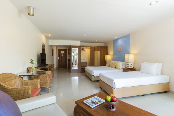 A hotel room with two beds, a sitting area with wicker furniture, a small table with a magazine and fruit, and a TV. - Bella Villa Cabana (Pattaya)