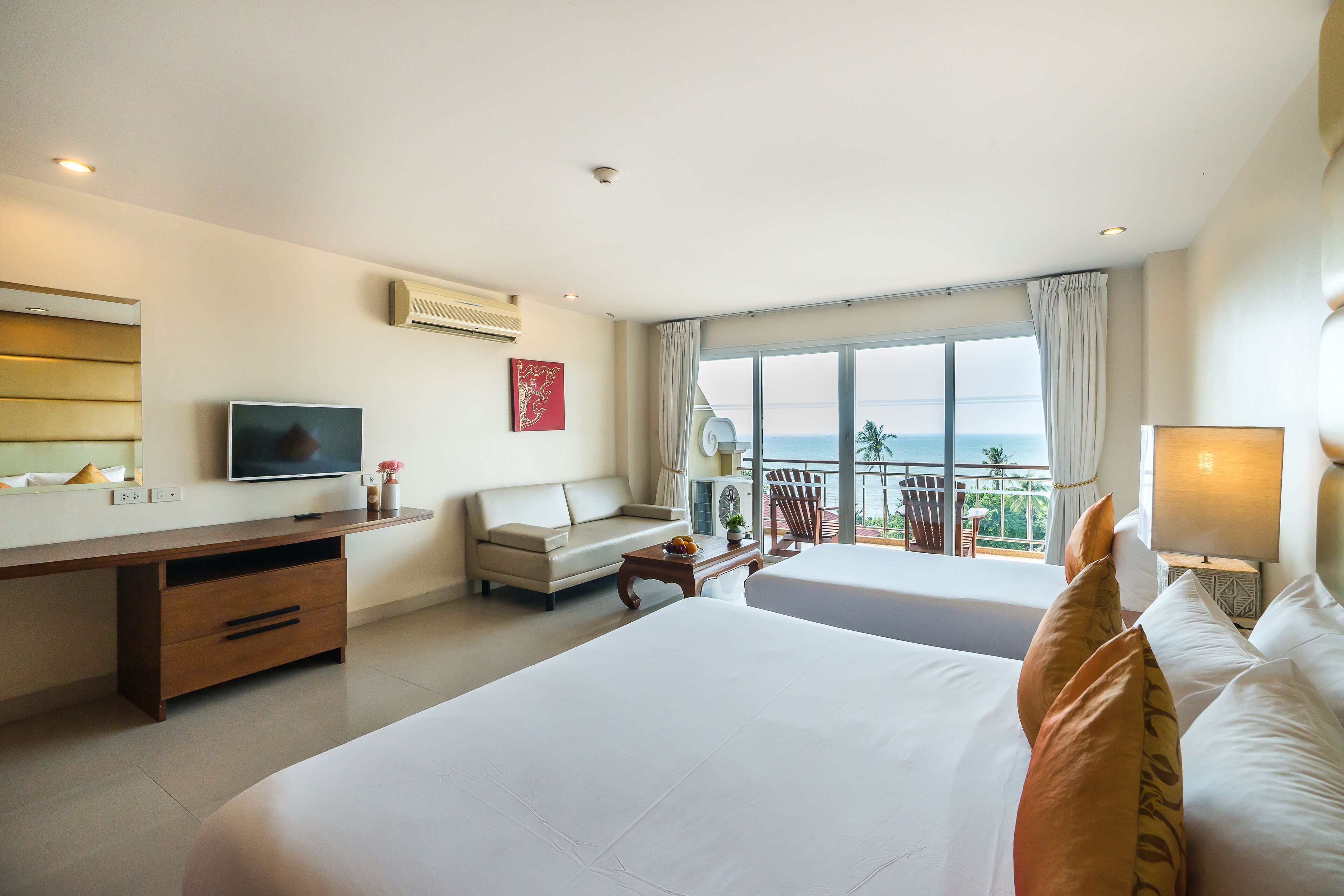 A hotel room with two beds, a desk, a TV, and a balcony with a view of the sea.
