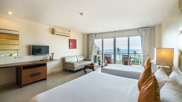 A hotel room with two beds, a desk, a TV, and a balcony with a view of the sea.