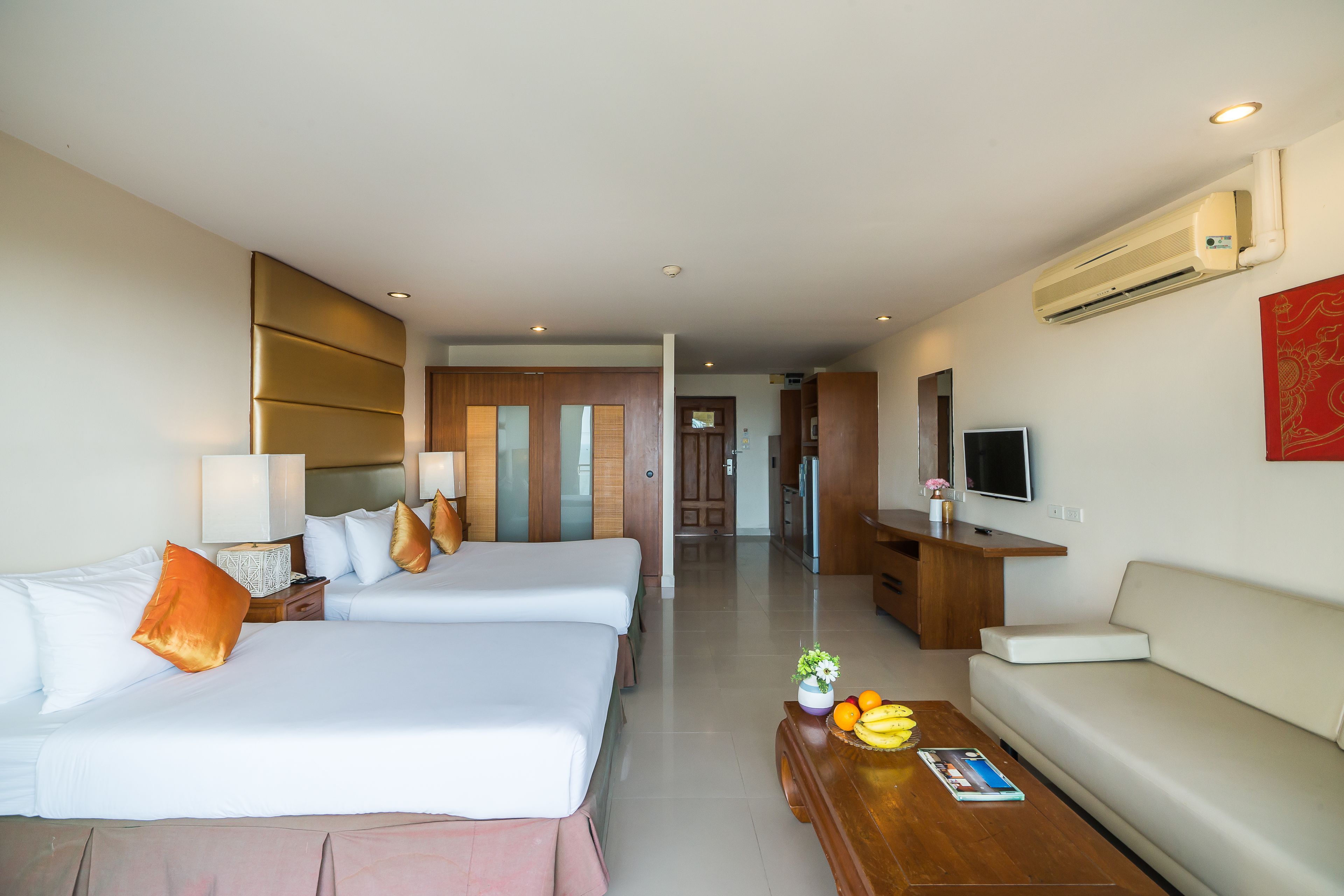 A hotel room with two beds, a sofa, a coffee table with fruits, a TV, and an air conditioning unit.