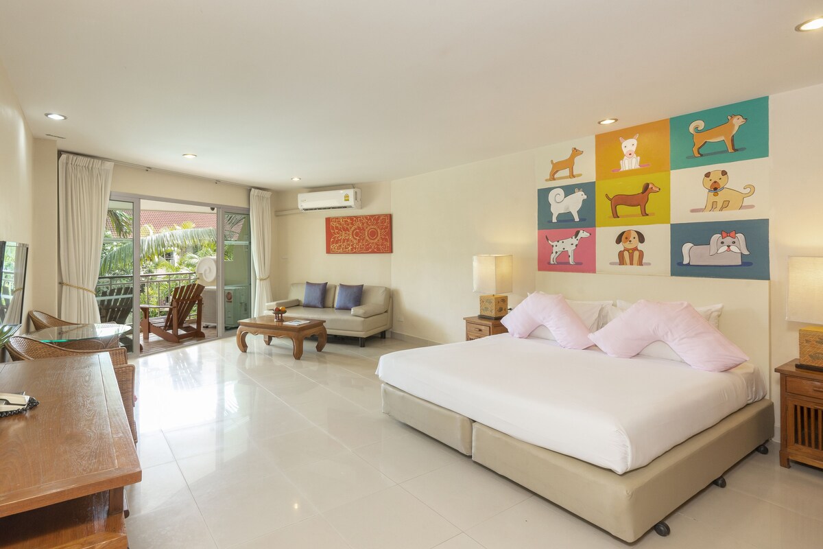 A spacious hotel room with a large bed, a sofa, a dining table, and a balcony with a view of greenery.