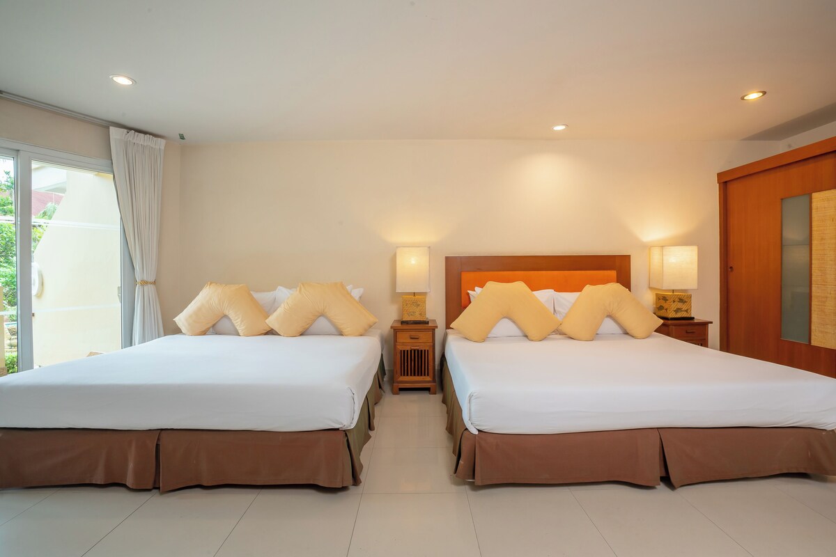 A hotel room with two beds, a wooden headboard, a nightstand with a lamp, and a view of the outdoors.