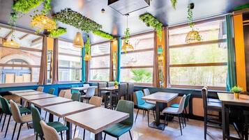 A restaurant with high ceilings, hanging plants, and large windows. Tables and chairs are arranged for dining.