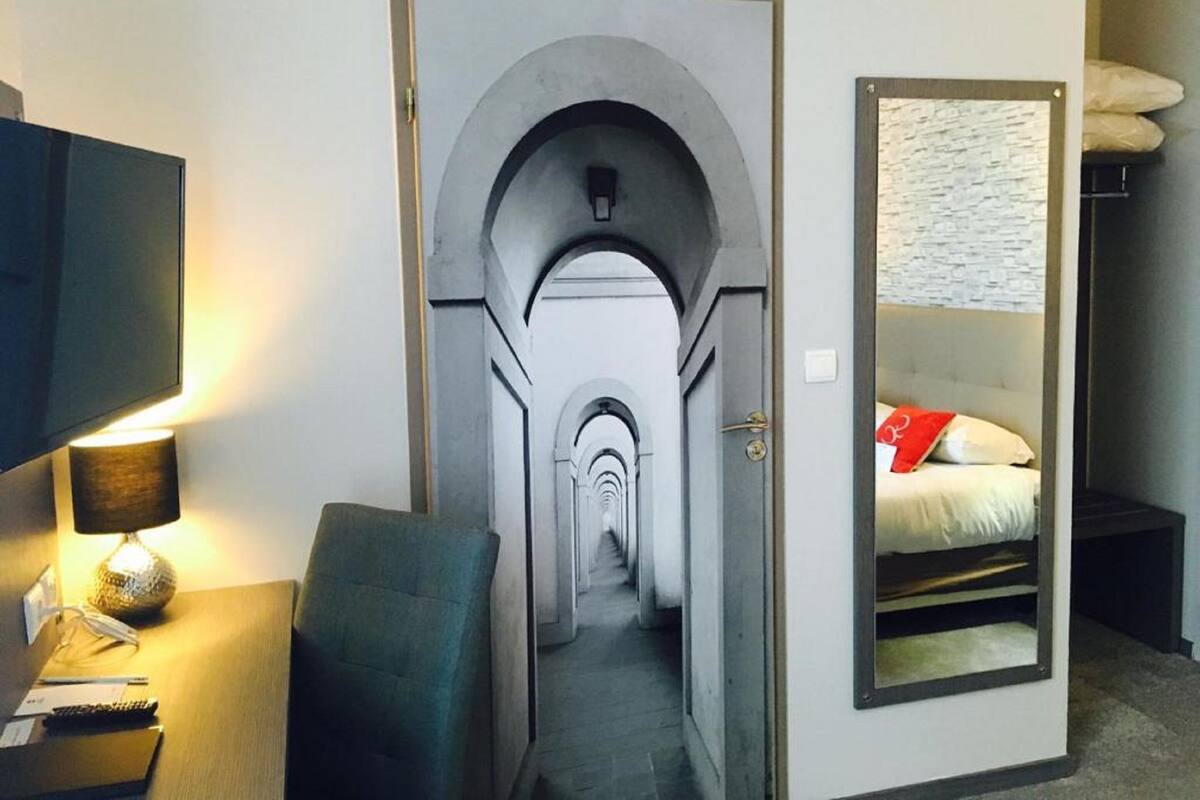 Ein Hotelzimmer mit einem schmalen Flur, der zu einem Bett, einem Schreibtisch mit Lampe und einem großen Spiegel führt, der das Bett reflektiert.