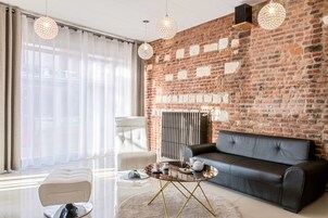 Una sala de estar moderna con pared de ladrillo, un sofá de cuero negro, un sillón blanco, una mesa de centro de vidrio y lámparas colgantes.