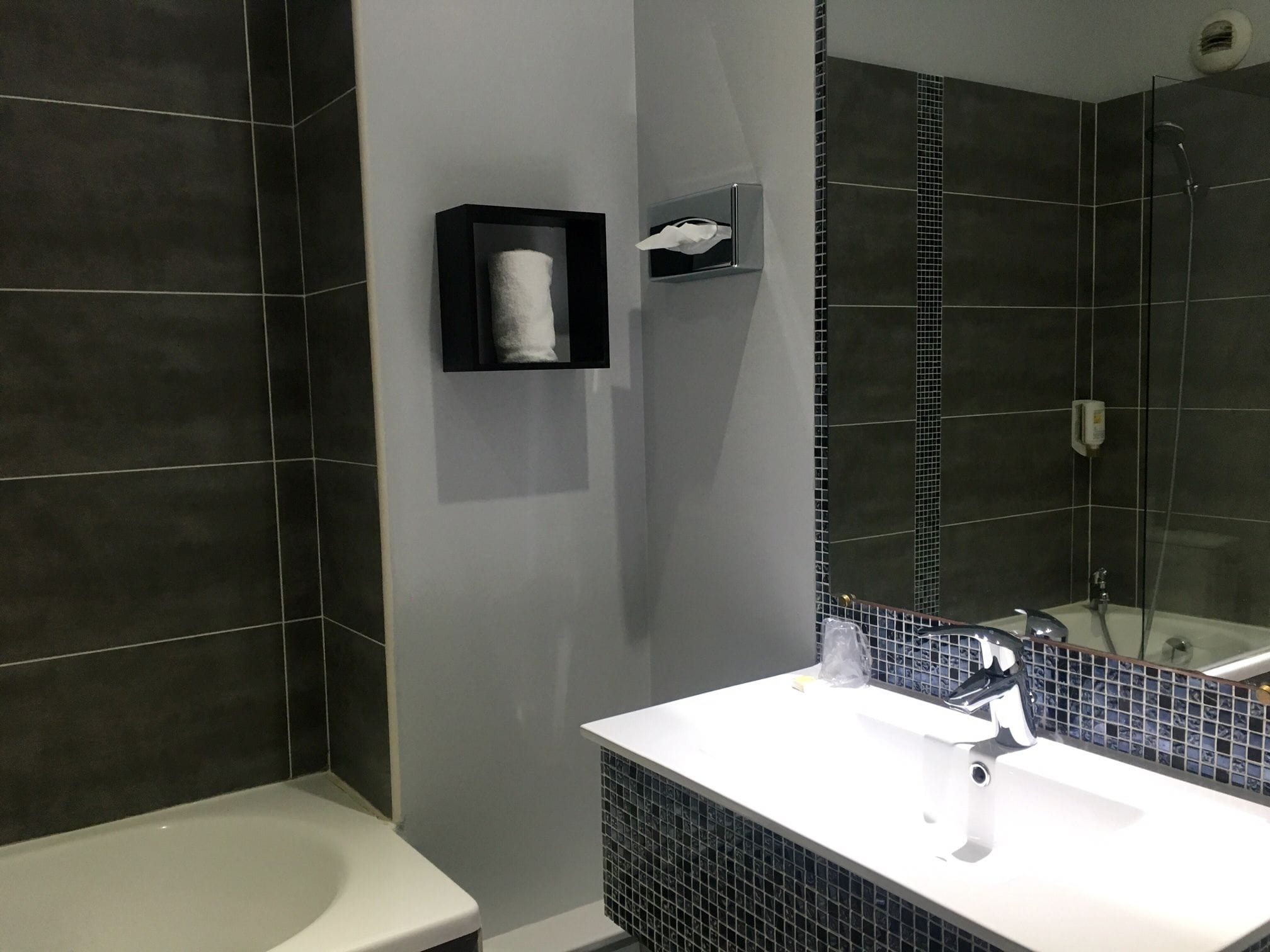 A modern bathroom with dark tiles, a white sink, and a glass shower enclosure.