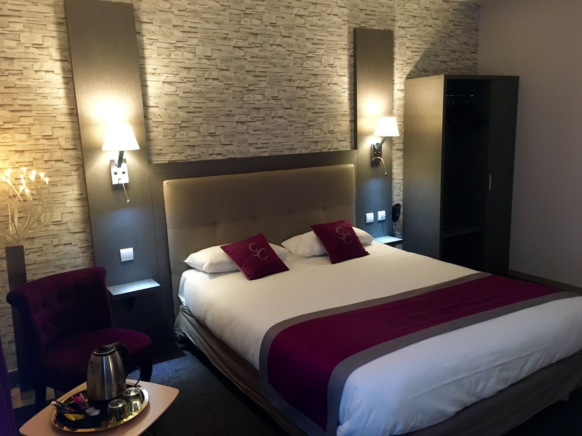 A hotel room with a large bed, two nightstands, a red armchair, a minibar, and a brick-patterned wall.