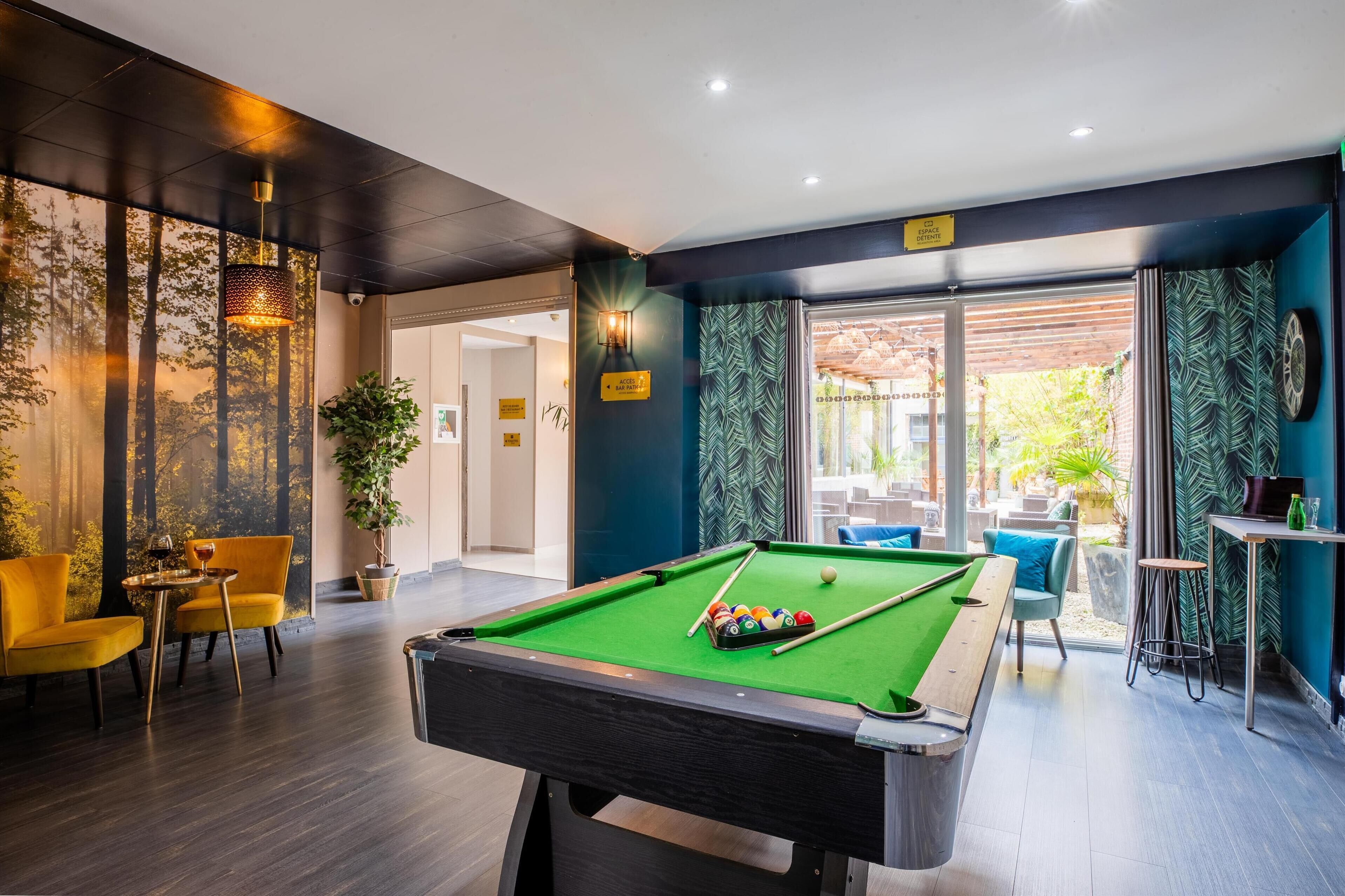 A pool table with a green felt surface, surrounded by yellow chairs and a small table with a glass of wine.