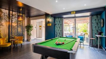 A pool table with a green felt surface, surrounded by yellow chairs and a small table with a glass of wine.