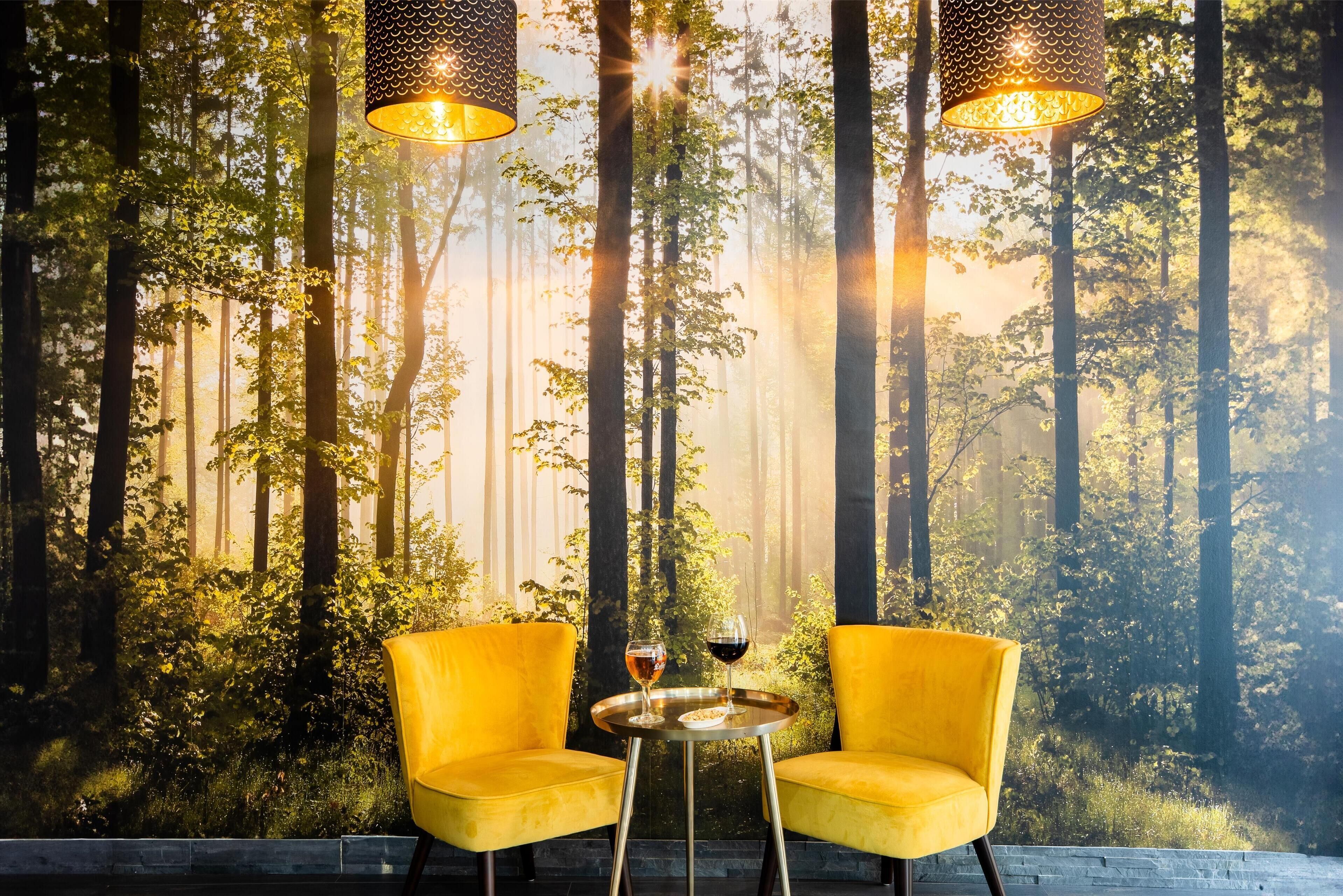 A room with a forest-themed wall mural, two yellow chairs, a small round table with a wine glass, and two hanging lamps.