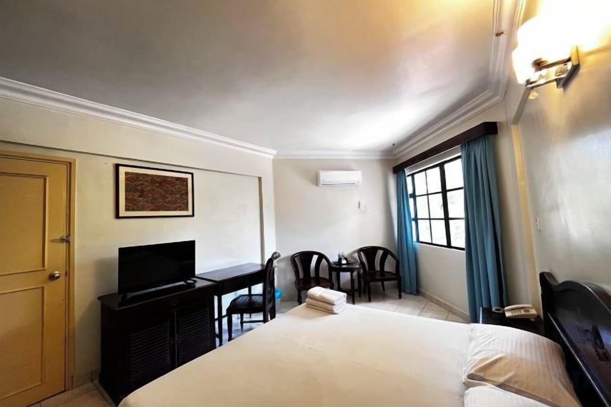 A hotel room with a bed, a desk with a television, a chair, and a window with curtains.