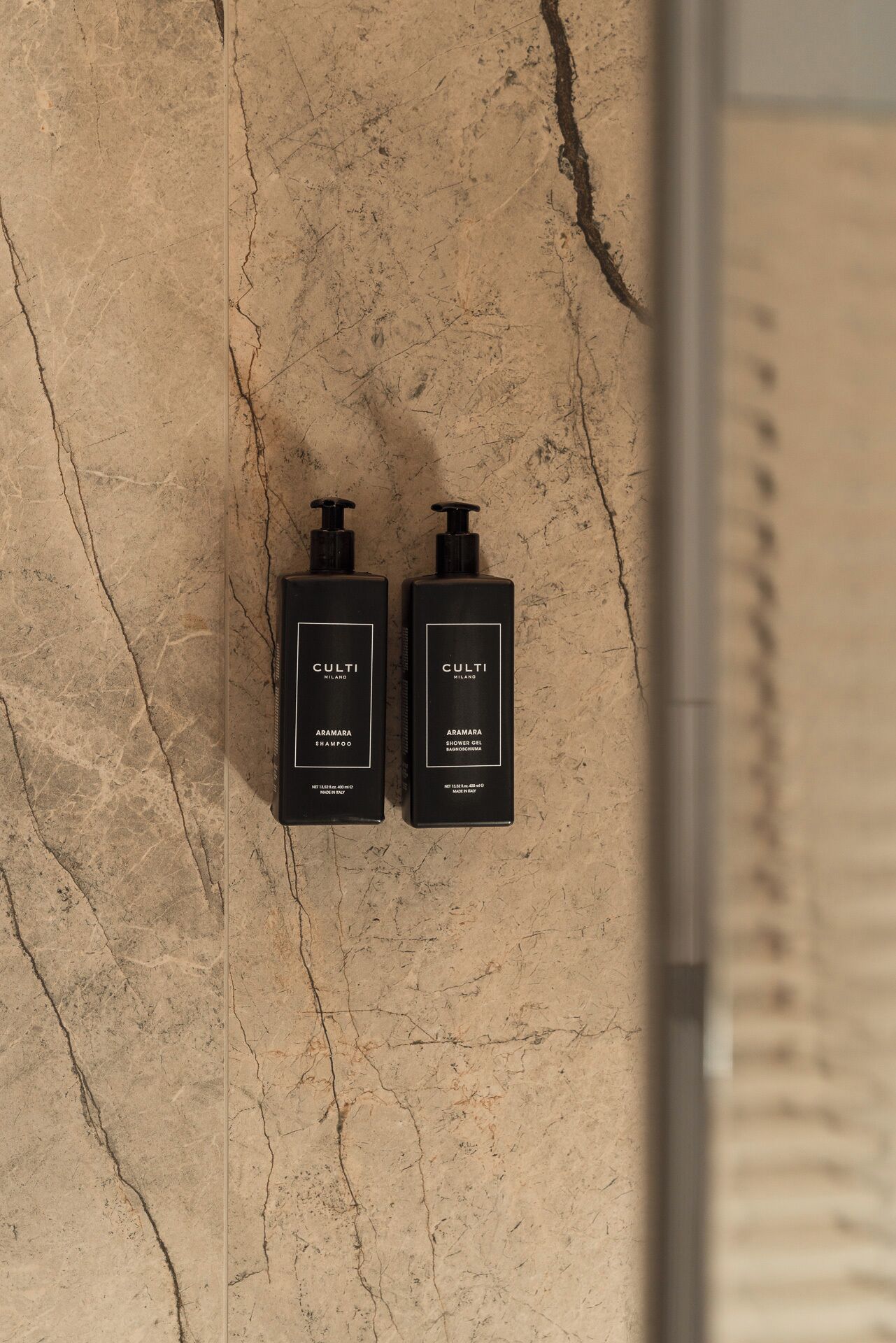 Two black bottles of Culti shampoo and shower gel on a textured beige wall.