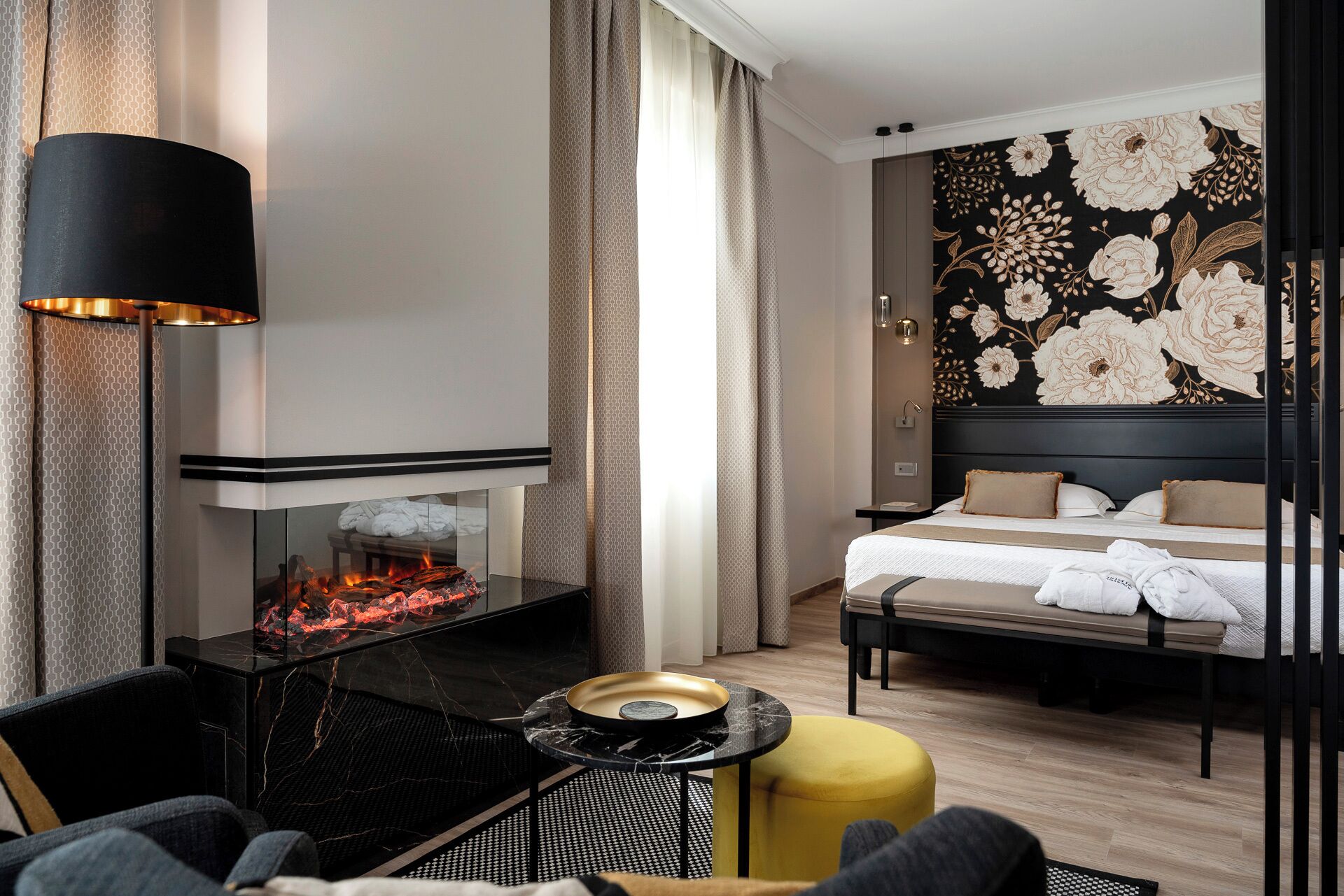 Une chambre d’hôtel moderne avec une cheminée, un lit avec du linge de maison blanc, un pouf jaune et un grand décor mural floral.