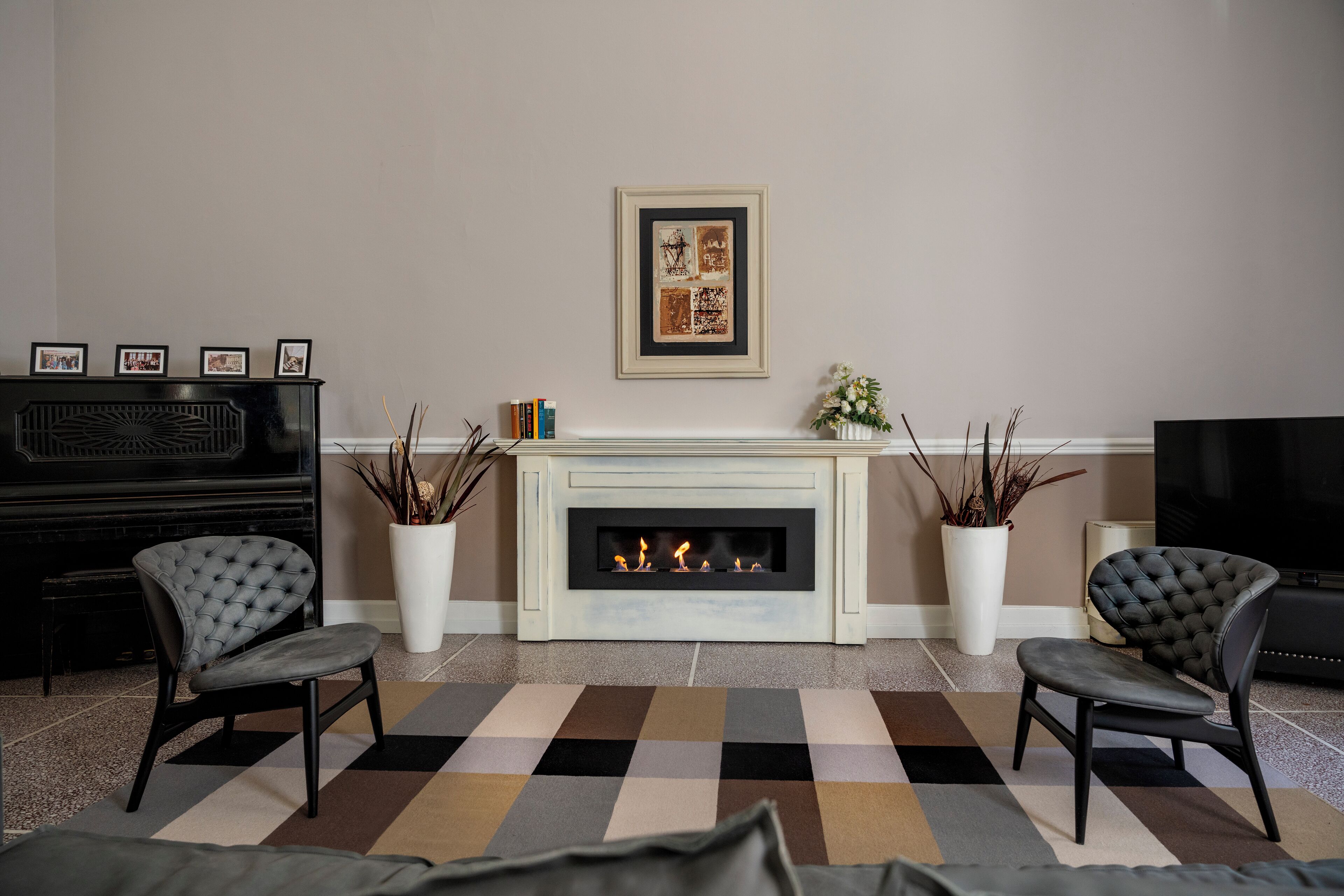Ein Wohnzimmer mit Kamin, zwei Sesseln, einem Klavier und einem gerahmten Kunstwerk an der Wand.