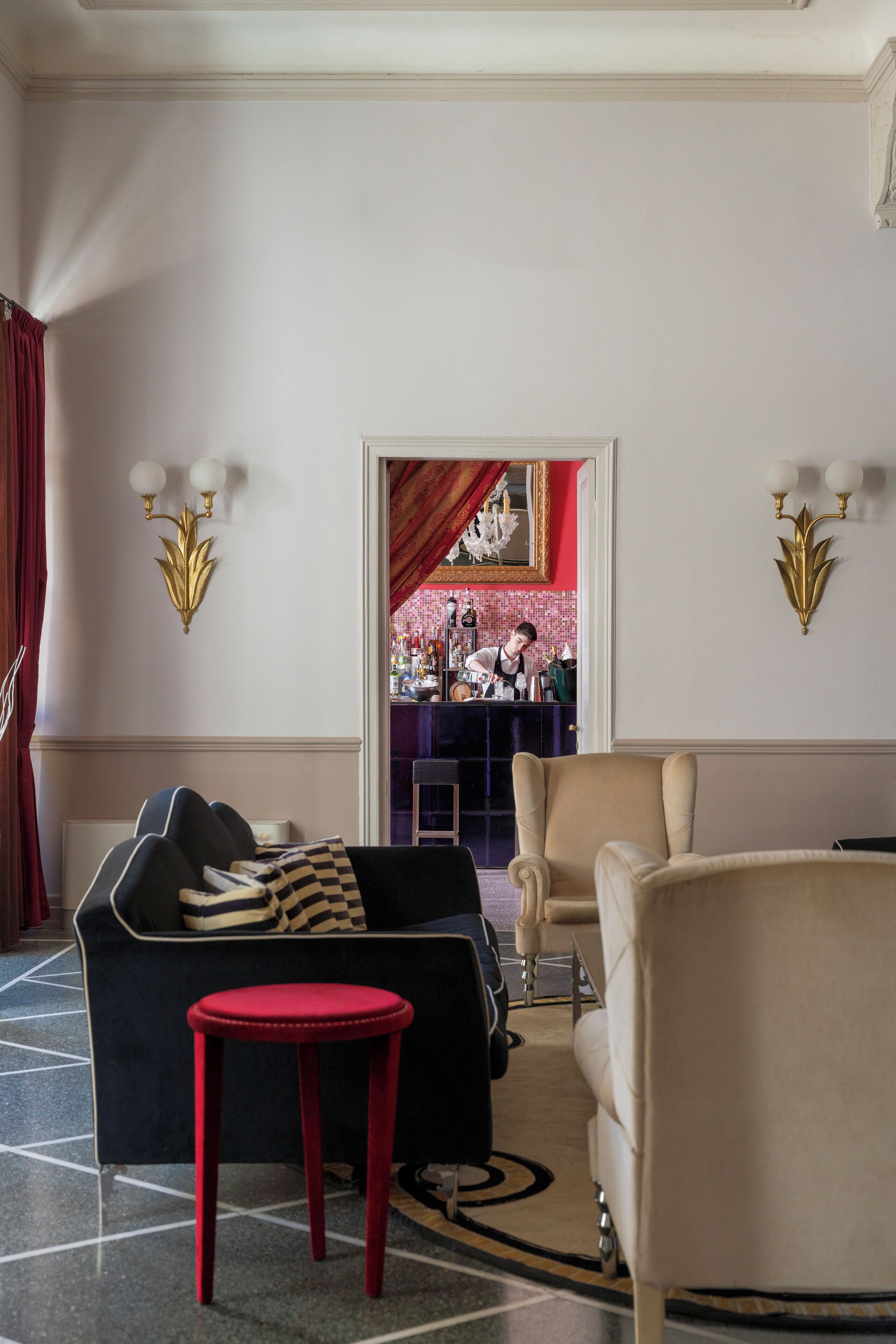 A hotel lobby with a black sofa, a red stool, a beige armchair, and a bar area in the background.
