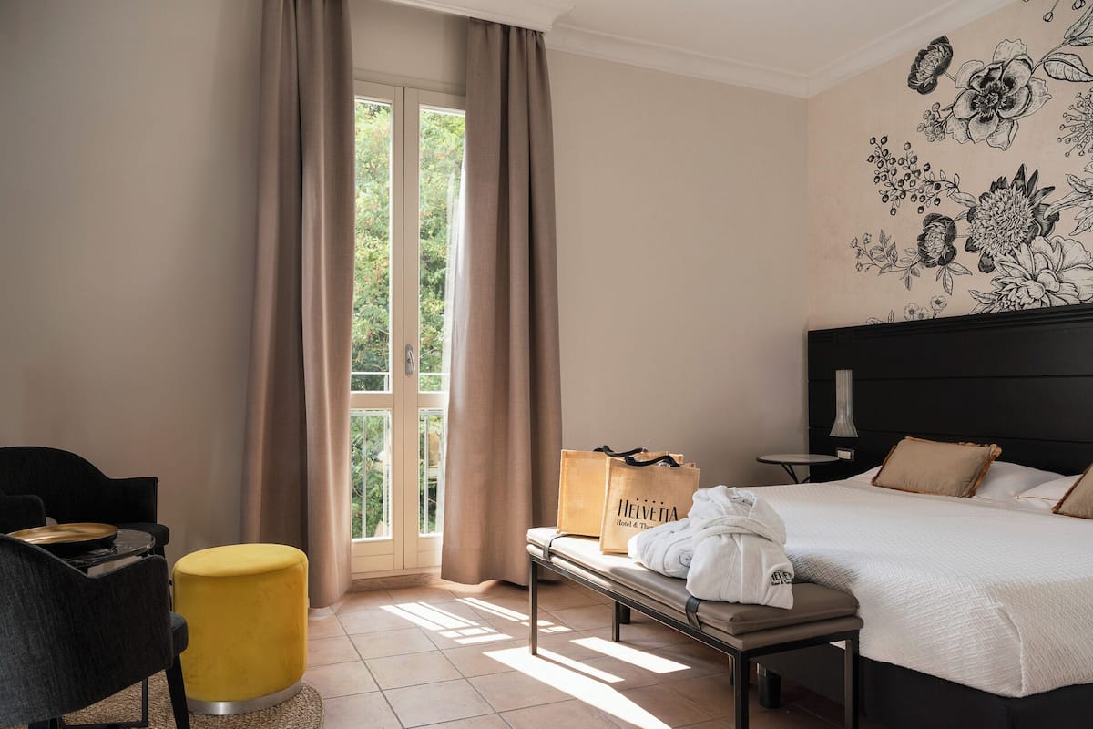 A hotel room with a bed, a bench, a chair, a table, and a window with curtains.