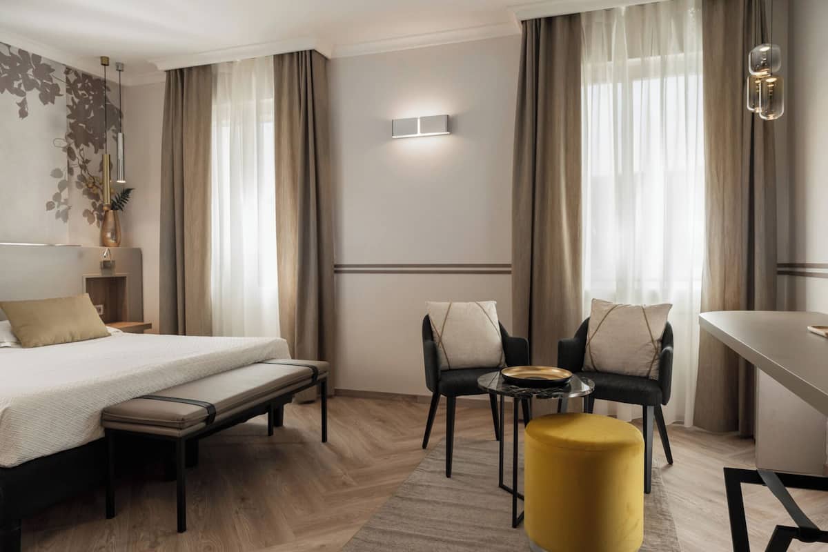 A hotel room with a bed, a bench, two chairs, a small table, a yellow ottoman, and a wall-mounted light fixture.