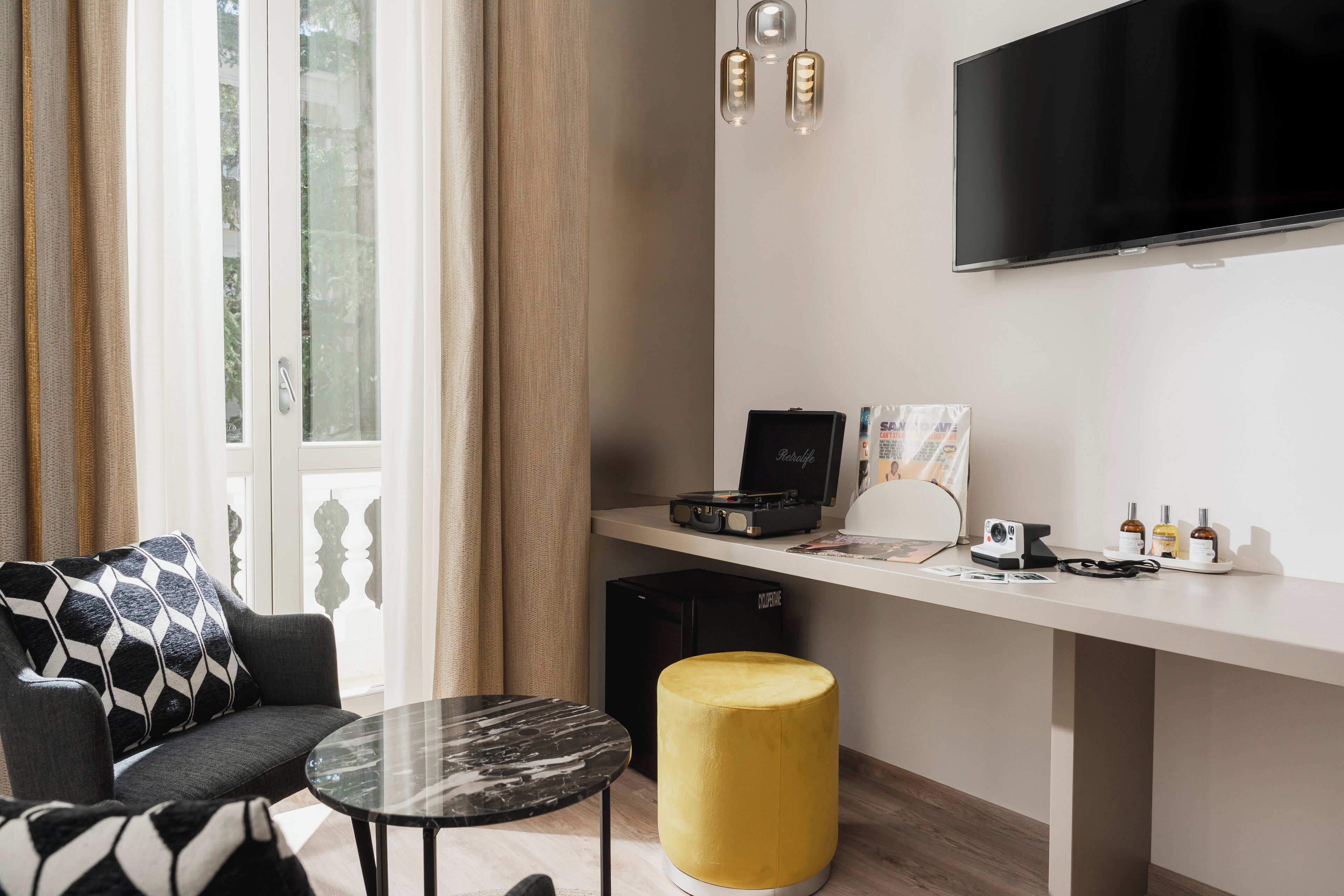 Un soggiorno moderno con un divano, un poggiapiedi giallo rotondo, un tavolino da caffè in vetro, una TV a schermo piatto, un giradischi e una piccola scrivania con un computer e alcuni oggetti sopra.